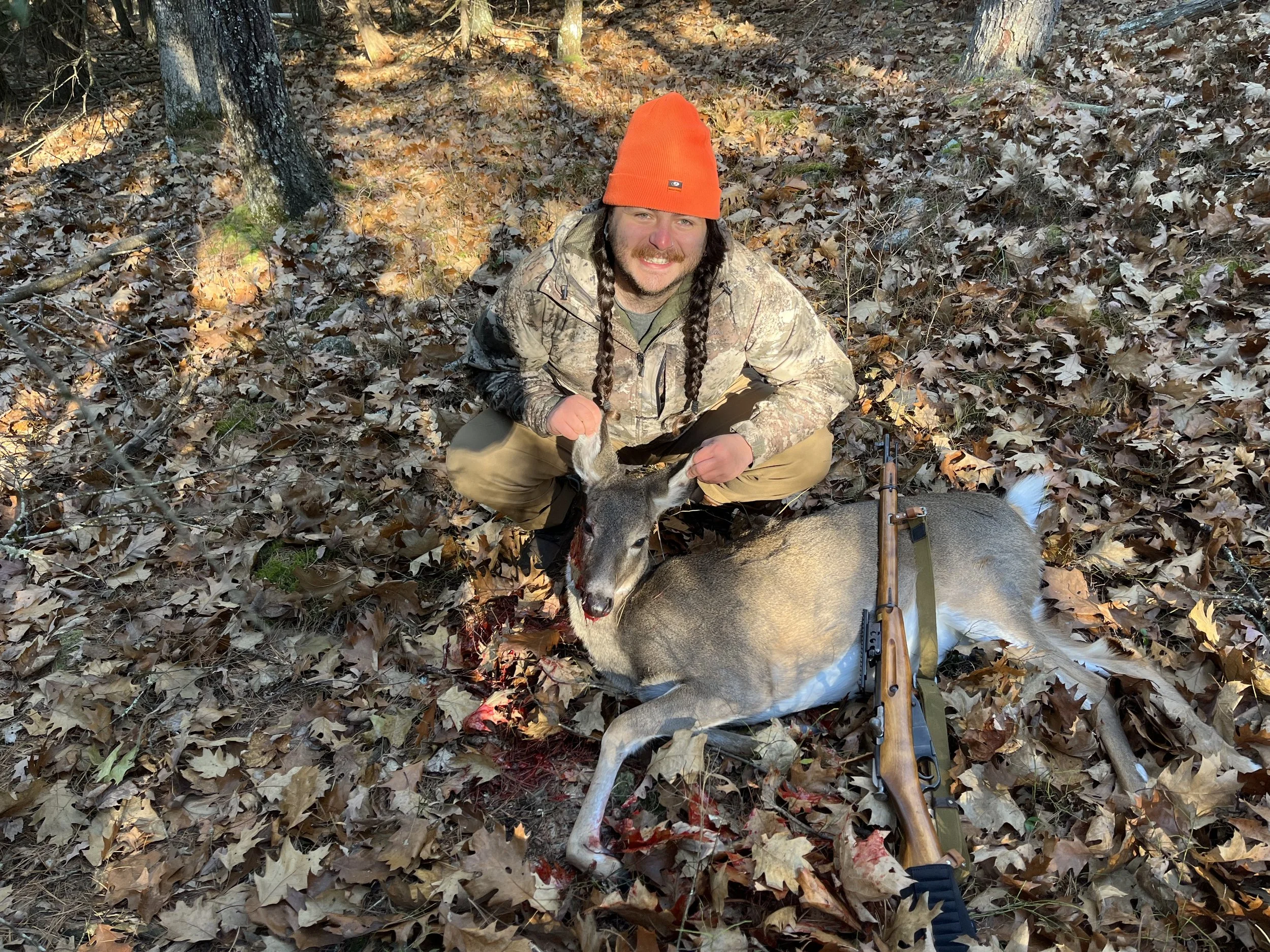 A man in camouflage clothing and an orange beanie squats in a leaf-covered forest, holding a deer by its antlers, with a rifle resting on the ground beside the deer.