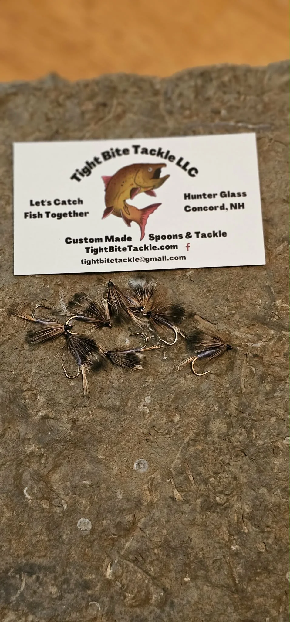 Close-up of several fishing flies with a business card from Tight Bite Tackle LLC, featuring a fish logo, contact information, and a slogan 'Let's Catch Fish Together' on a textured surface.