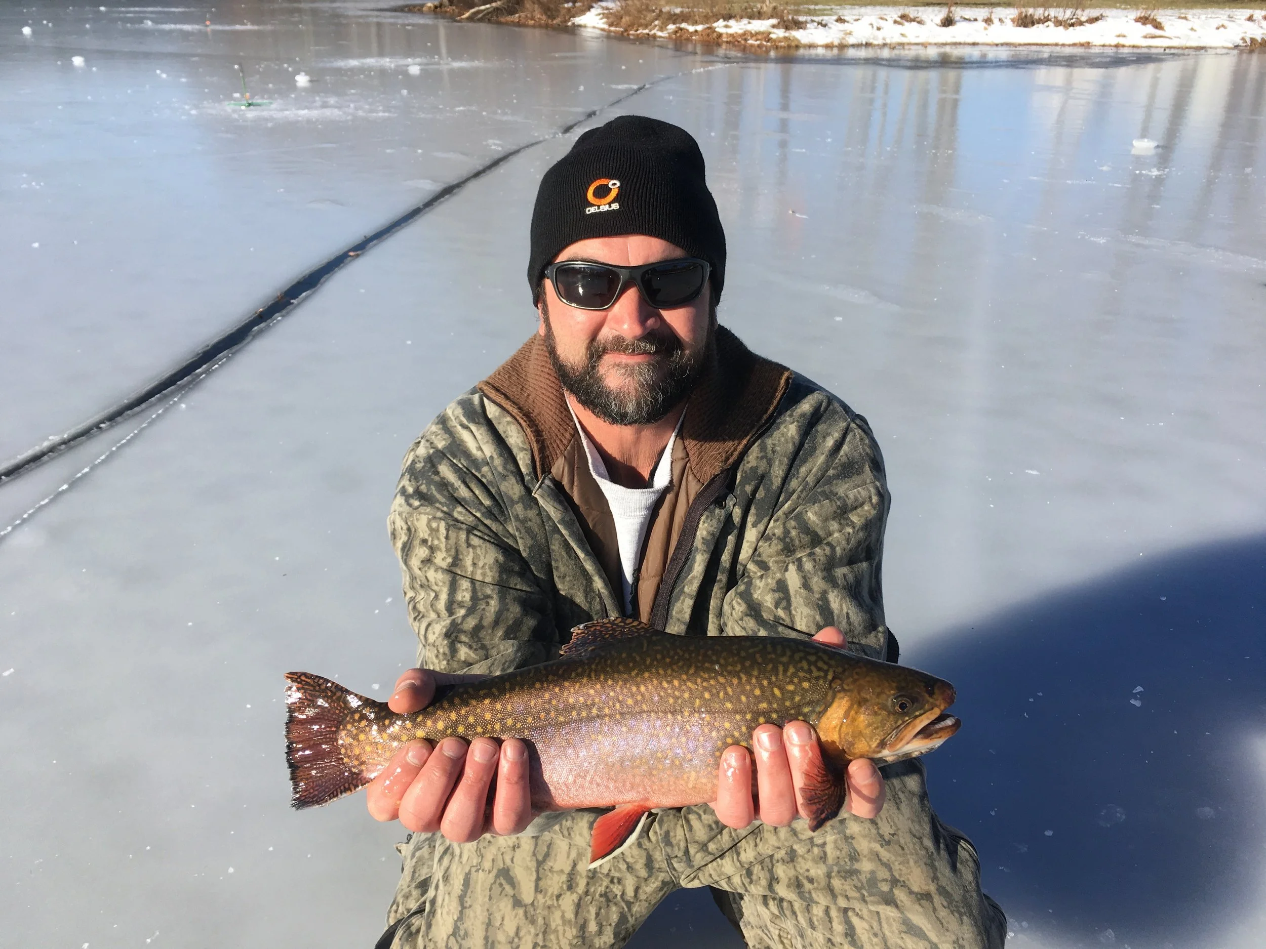 A man wearing sunglasses, a black beanie, and camouflage clothing kneels on ice, holding a fish with both hands, on a frozen body of water during the daytime.