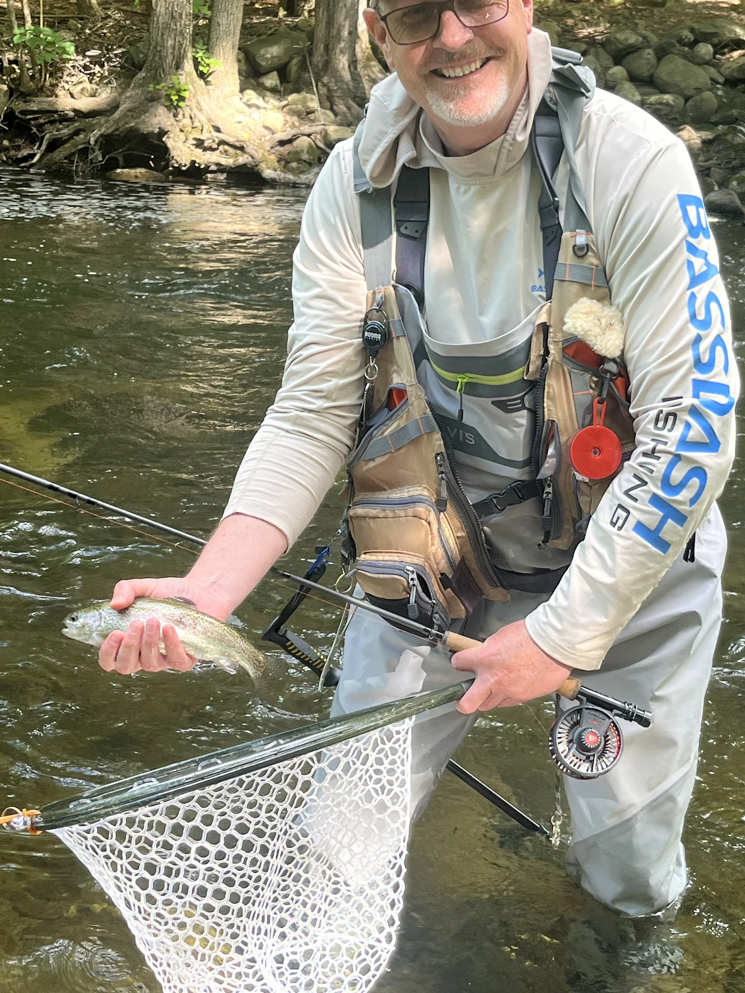 A man fishing in a river holding a small fish over a net. He is smiling, wearing waders and a fishing vest, with trees and rocks in the background.