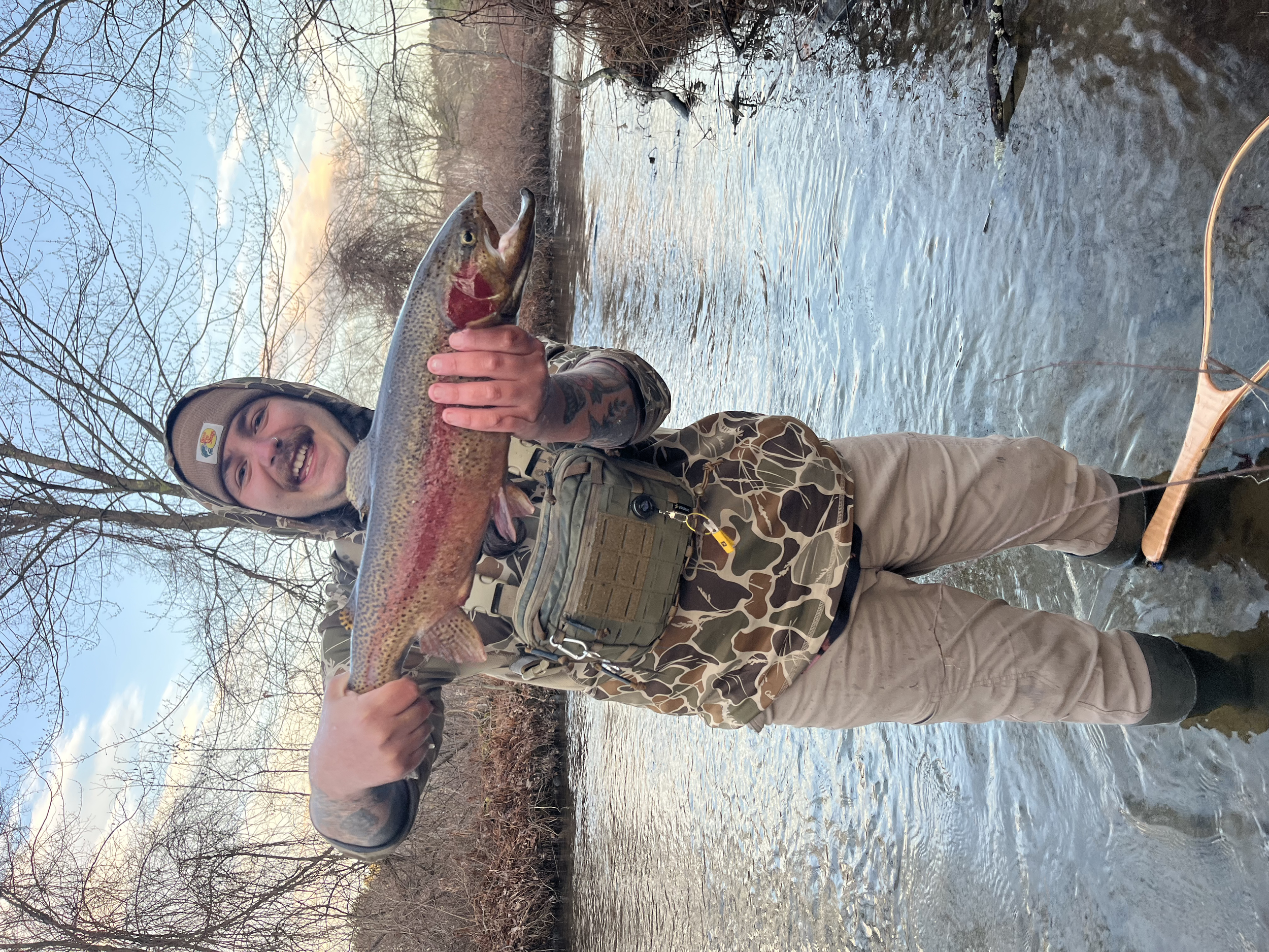 Man standing in a river holding a large rainbow trout fish he caught, smiling at the camera, winter trees in the background, wearing camouflage jacket, beige pants, and a beanie.