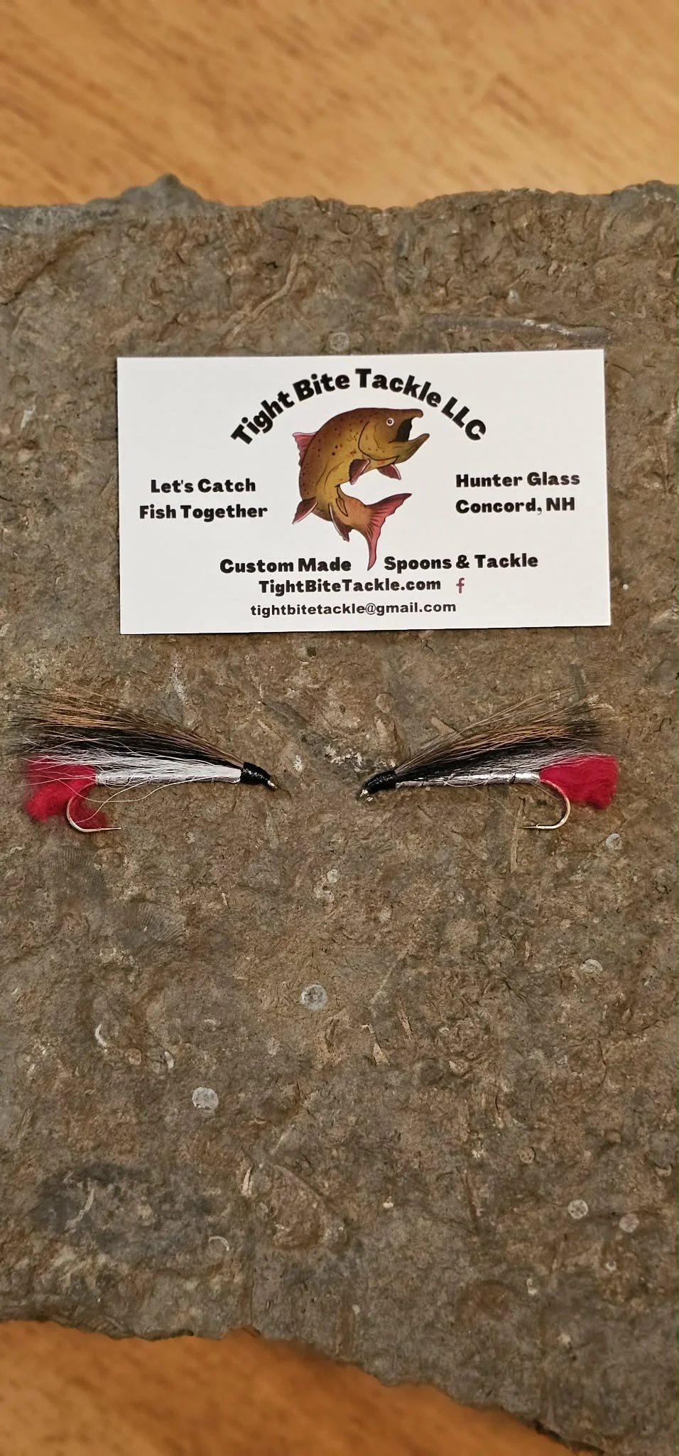 Two fishing flies with black, white, red, and pink feathers on a brown, textured surface. A business card from Tight Bite Tackle LLC with a cartoon fish logo and contact information is placed above the flies.