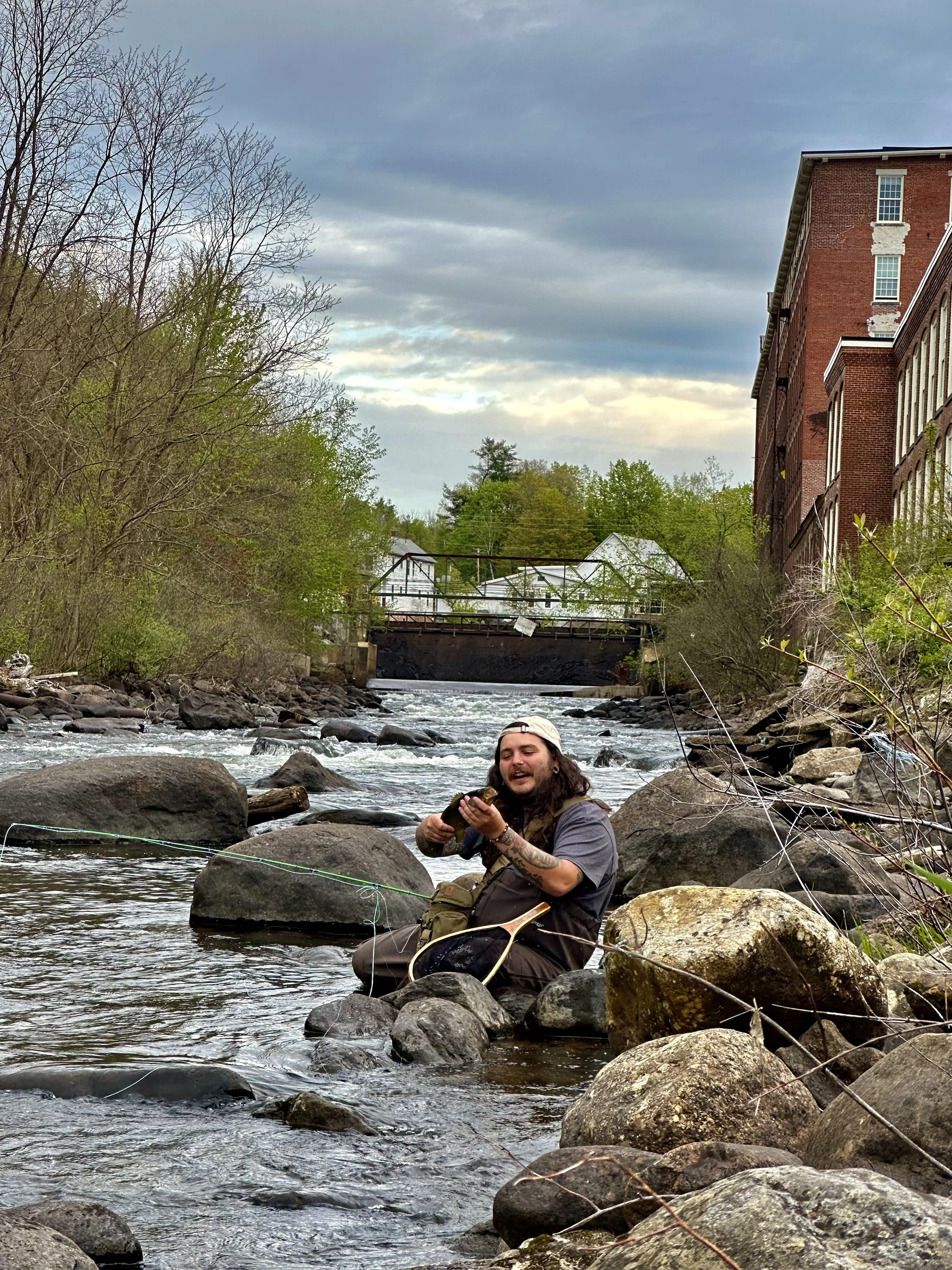A man fishing by a river with rocks, trees, a bridge, and buildings in the background.