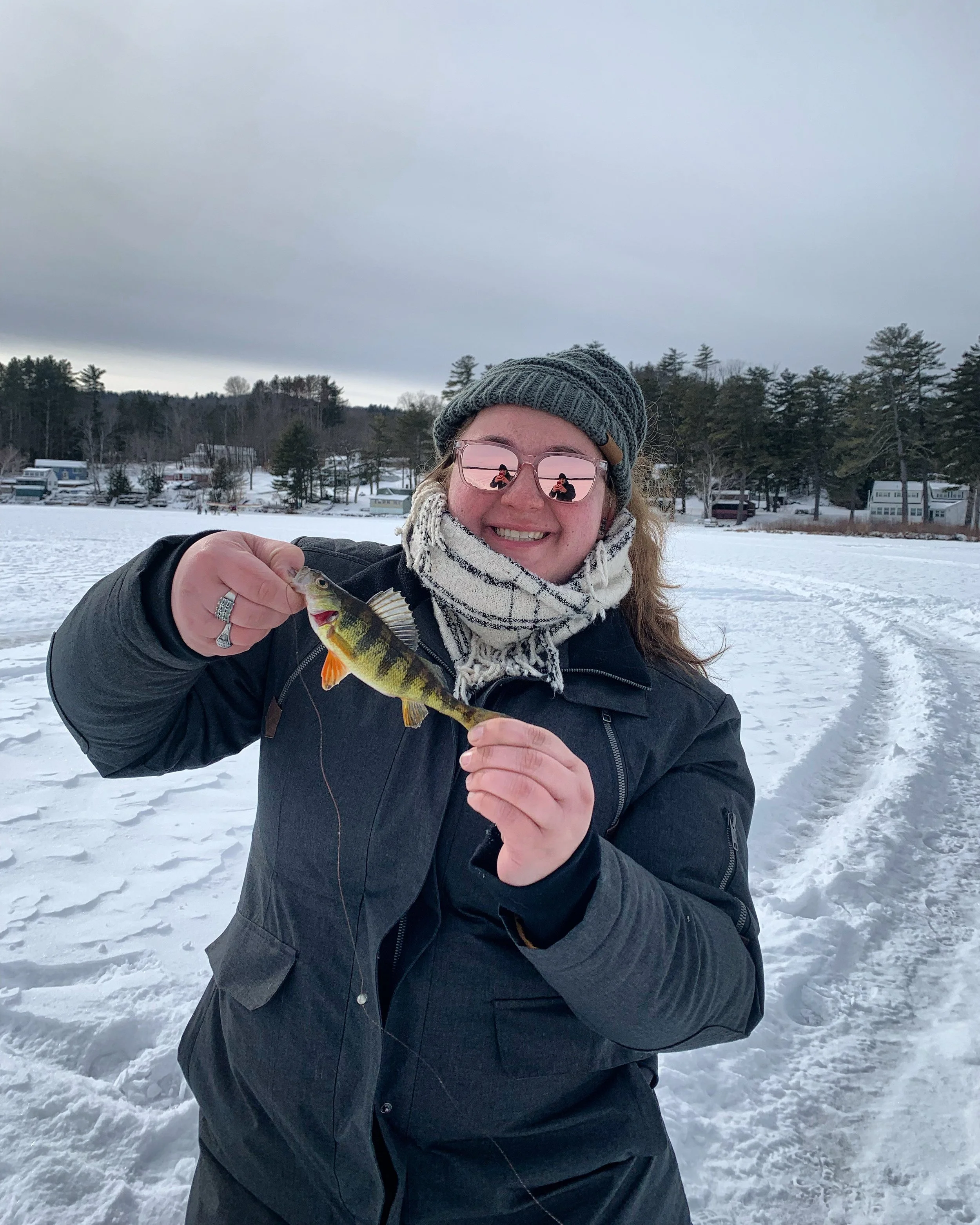 Woman in winter clothing holding a fish outdoors on snow-covered ground during overcast weather.
