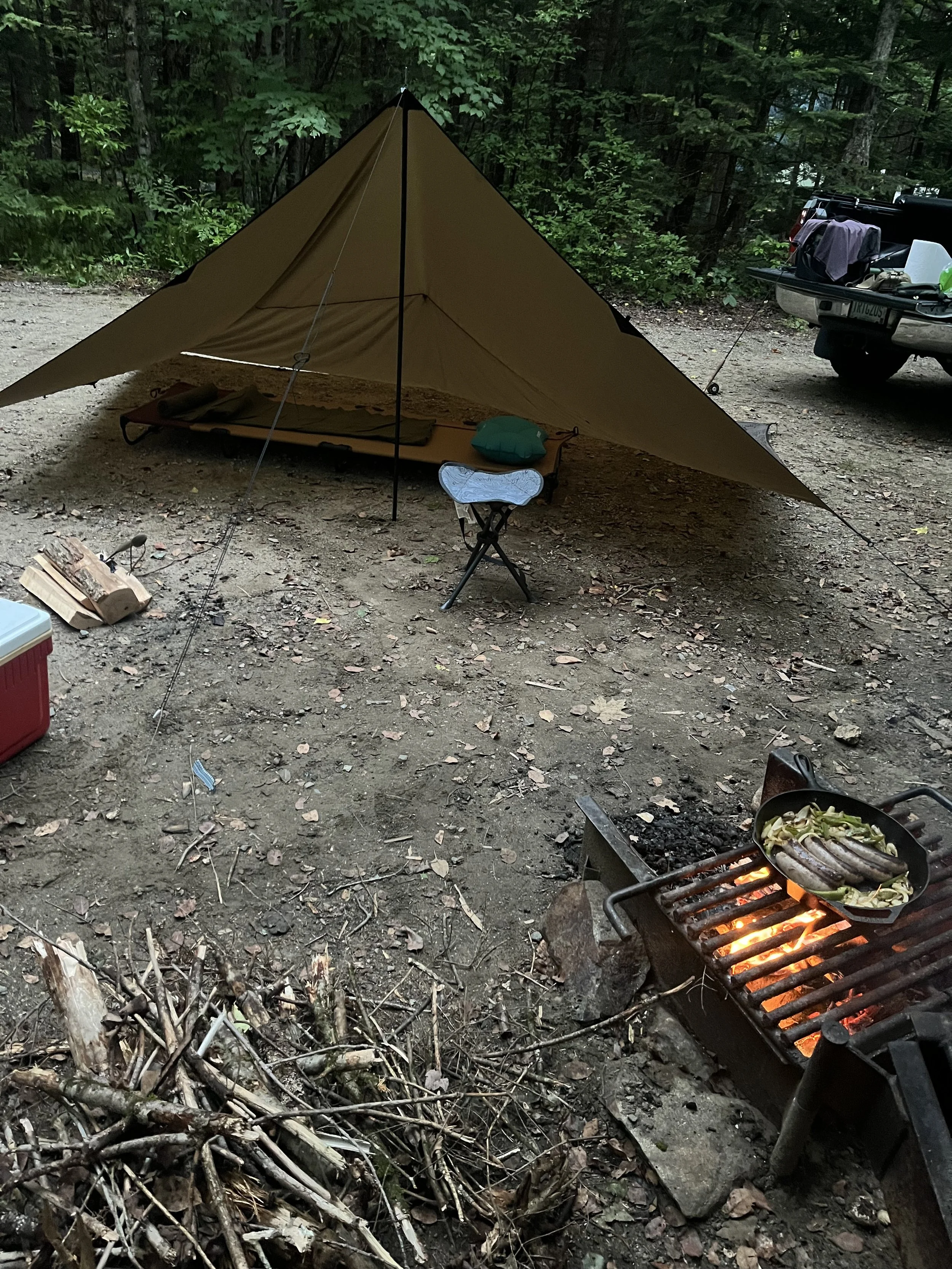 A campsite in a wooded area with a tan tent, a small folding stool with a map on it, a fire pit with sausages and vegetables cooking on a grill, a red cooler, a pile of firewood, and a pickup truck with luggage on the back.