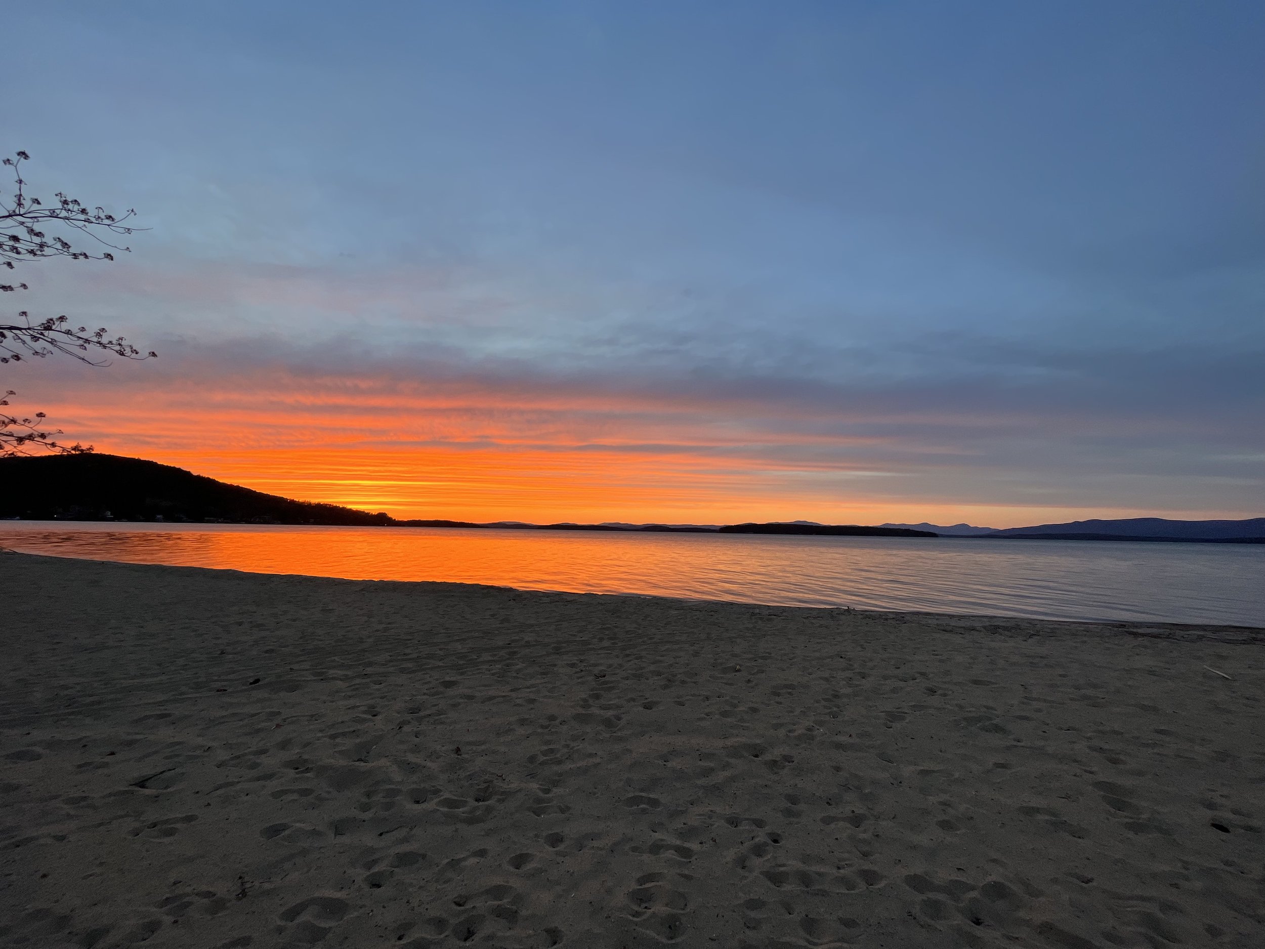 Beach at sunset with orange and blue sky over calm water and distant hills.