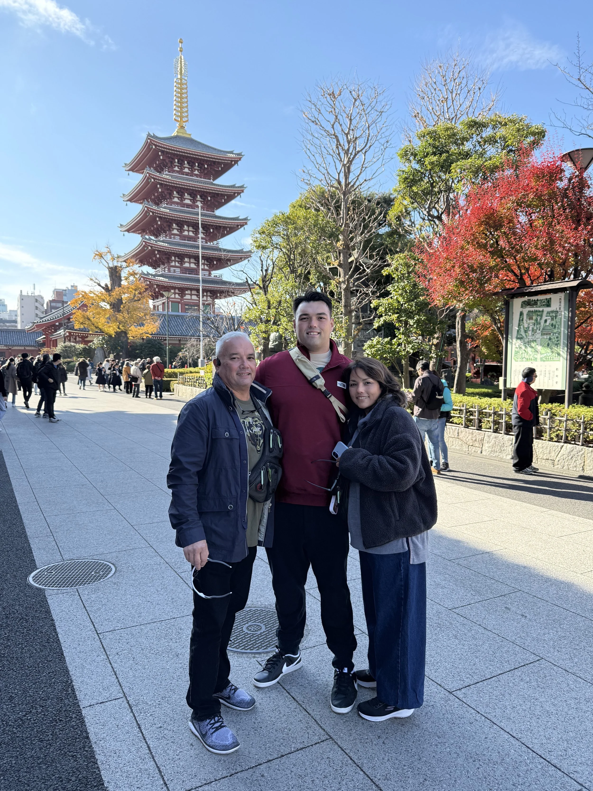 Three people standing in front of Senso-ji Temple in Tokyo, Japan, with colorful autumn trees and a clear blue sky in the background.