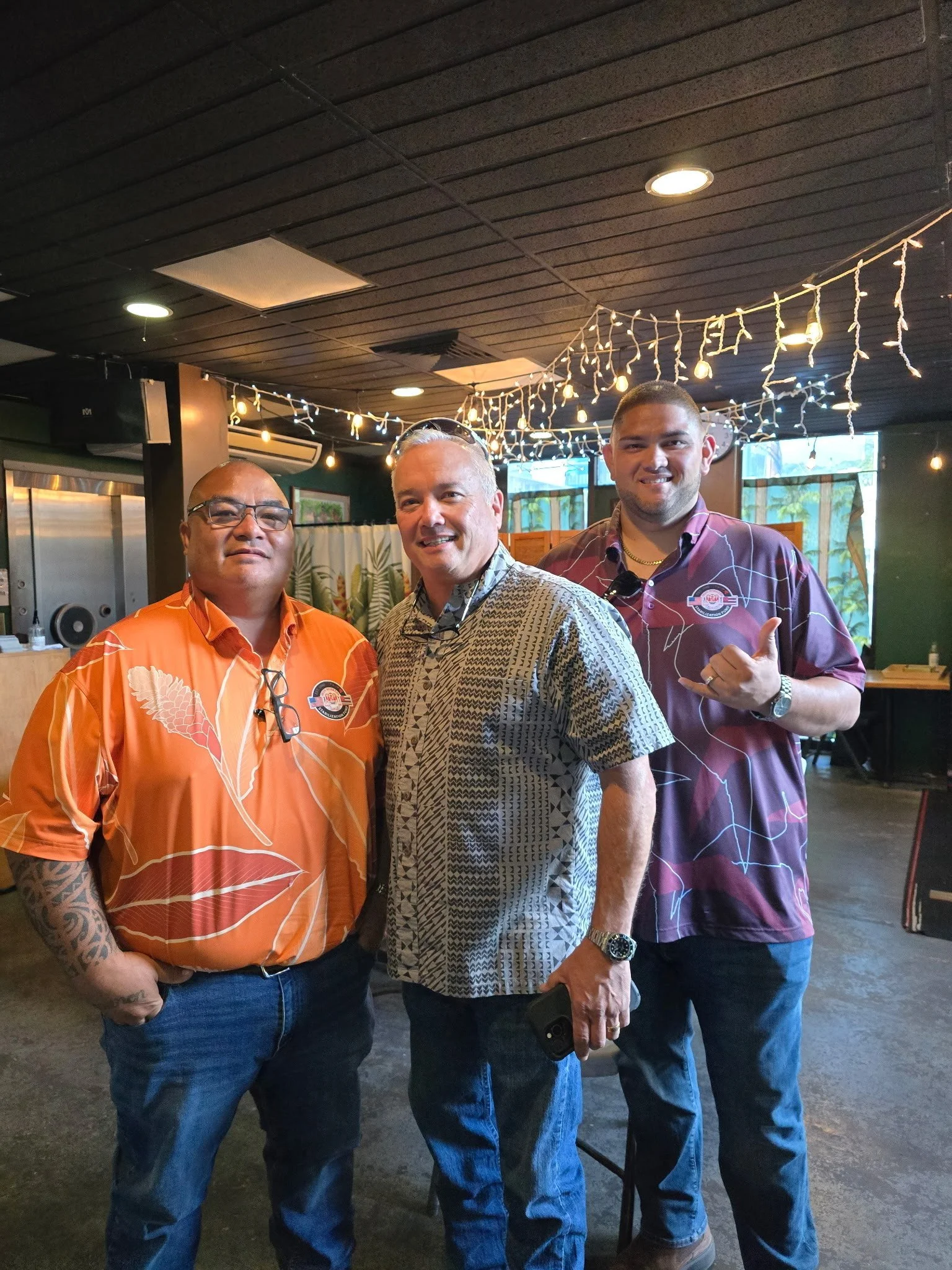 Three men posing together indoors, decorated with string lights hanging from the ceiling.