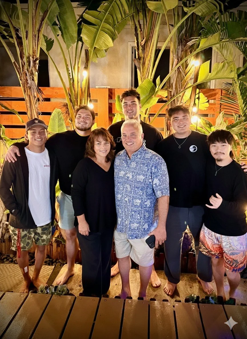 Group of six people, three women and three men, standing outdoors in front of large tropical plants with lights, smiling.