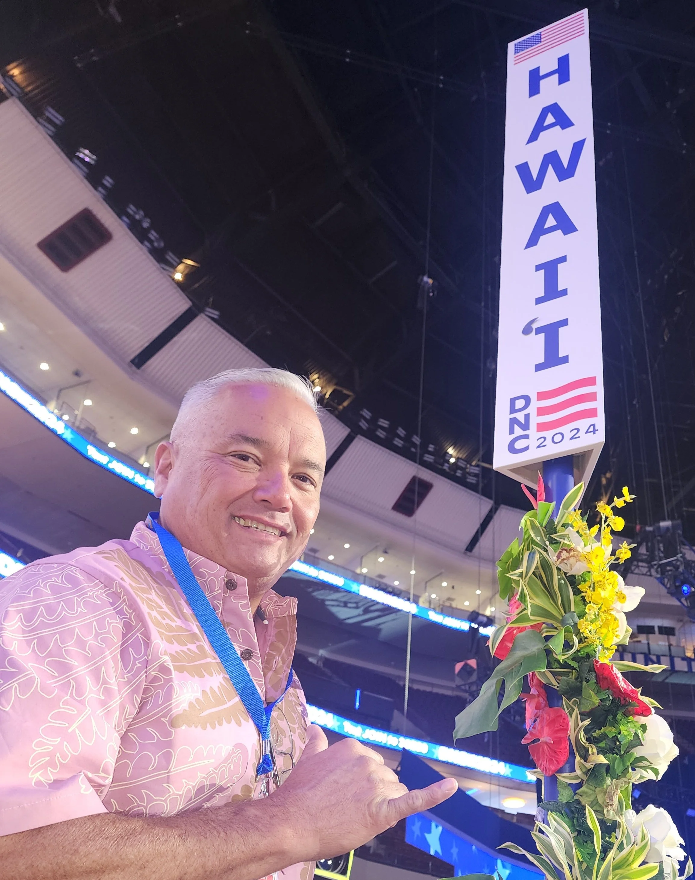 A smiling man in a pink patterned shirt with a blue lanyard, standing in front of a large sign that says 'HAWAII' with the American flag and 'DNC 2024' at a convention or event venue.