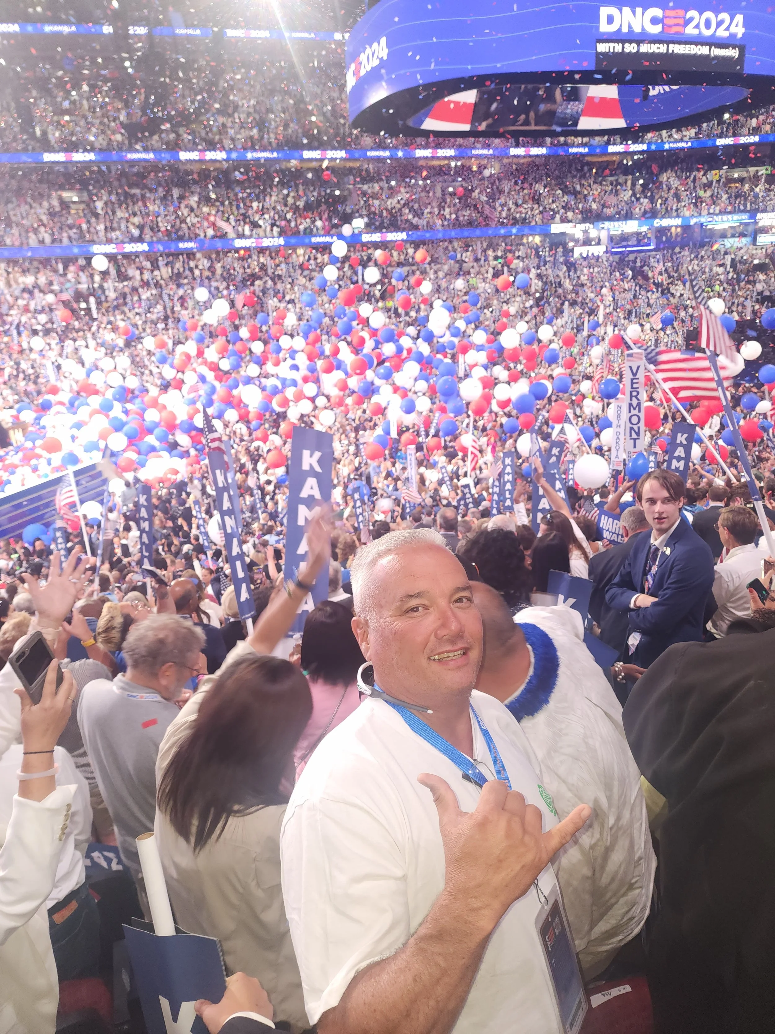 Crowd at a political event with balloons, flags, and banners, celebrating at a rally for DNC 2024, with a man in the foreground making a hand gesture and smiling