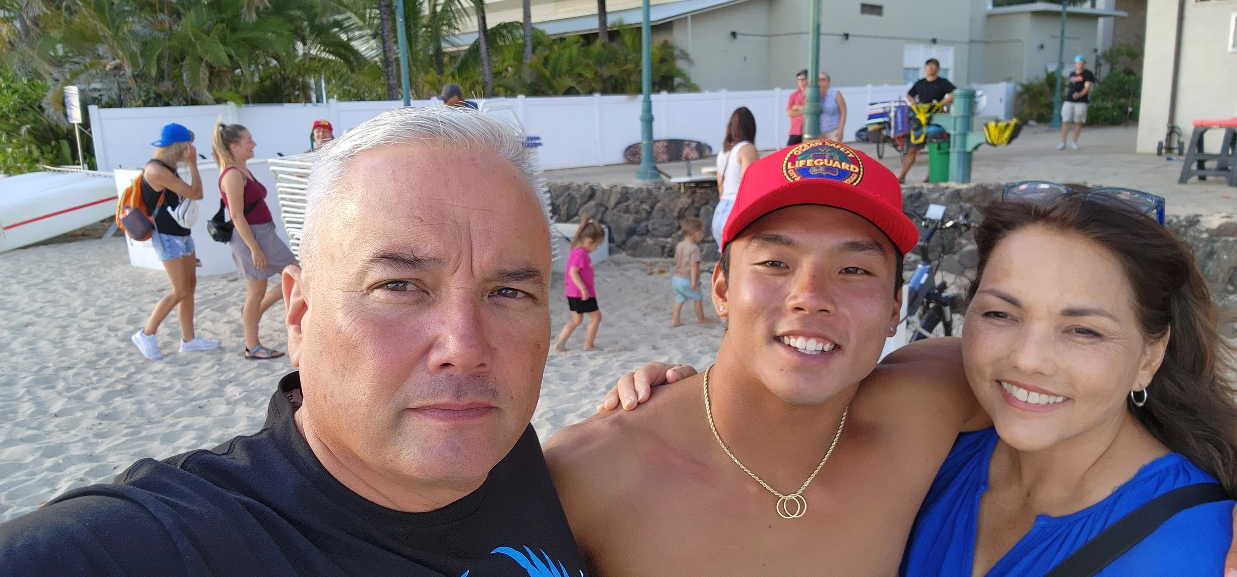 Three people taking a selfie on the beach near a white fence, with a beach area, children, and people in the background.