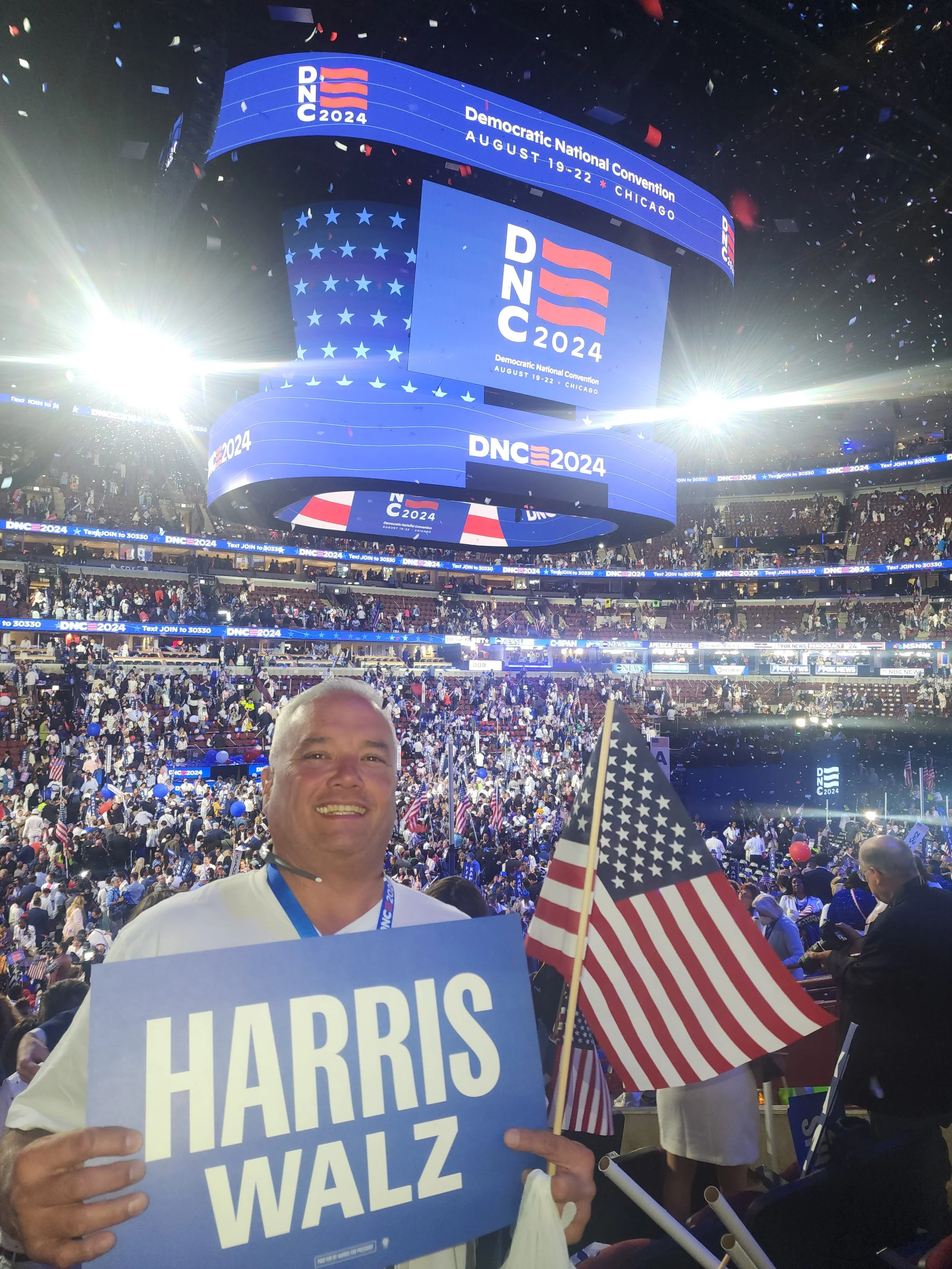 A person holding a sign that reads 'HARRIS WALZ' at the Democratic National Convention in Chicago, August 2024, with an American flag, surrounded by a large crowd and multiple flags, inside a brightly lit stadium with a large screen displaying the DN