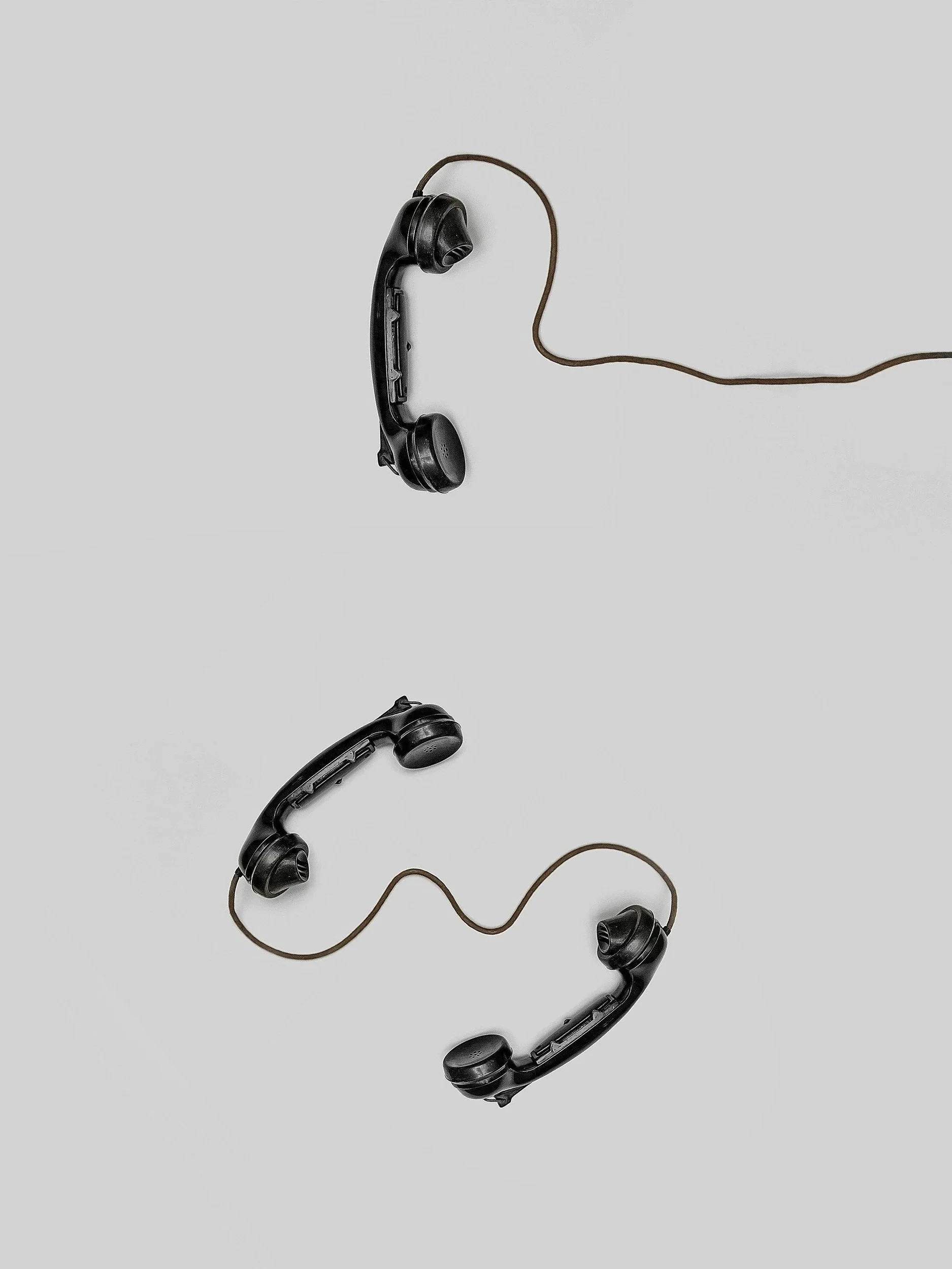 Two vintage black rotary phones connected by a single cord, suspended in mid-air against a plain gray background.