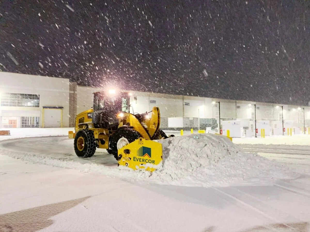 A yellow snowplow with the logo of Evercor clearing snow outside of a warehouse during a snowfall at night.
