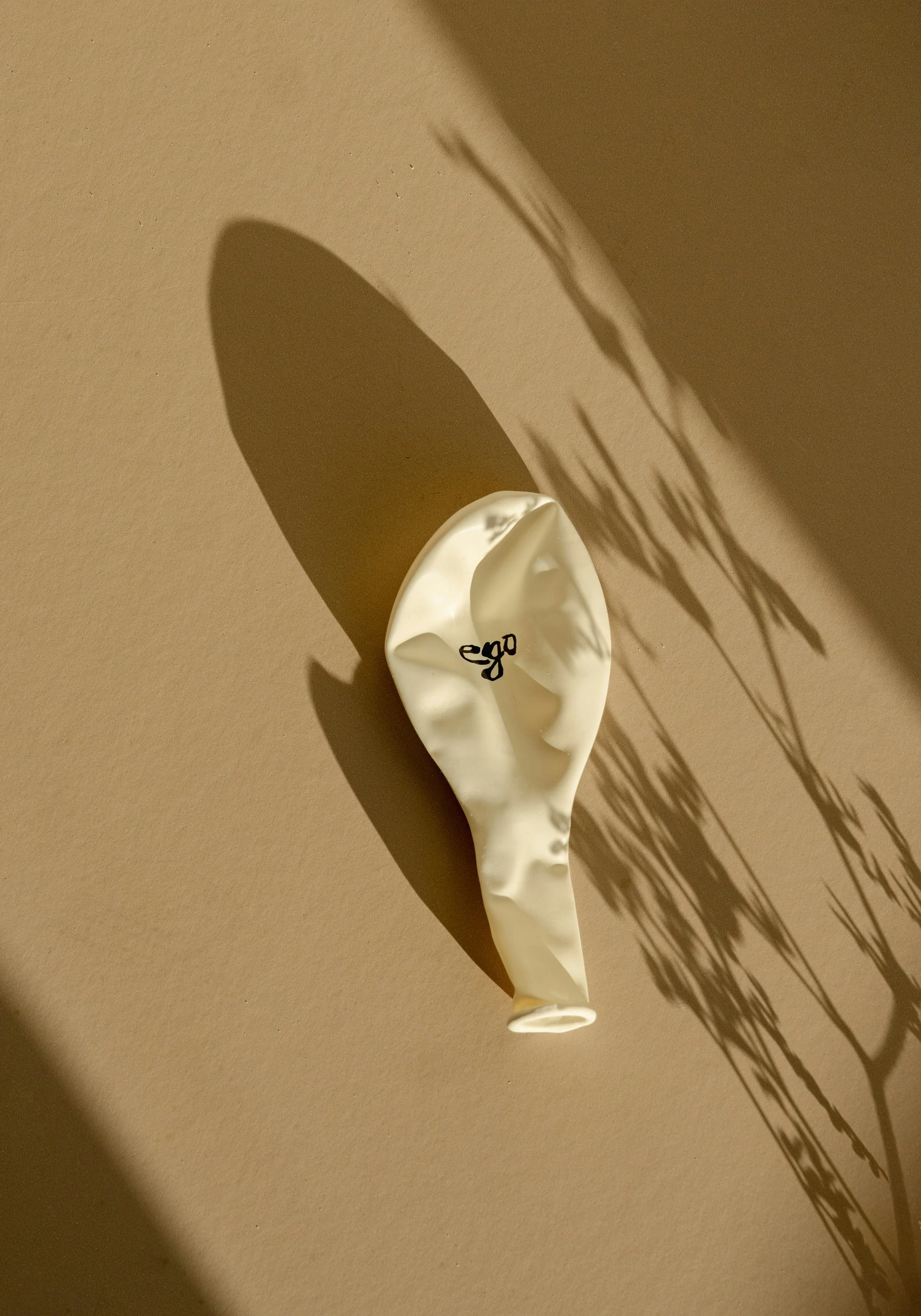 A crumpled white balloon with the word 'ego' written on it in black, casting a shadow on a beige wall.