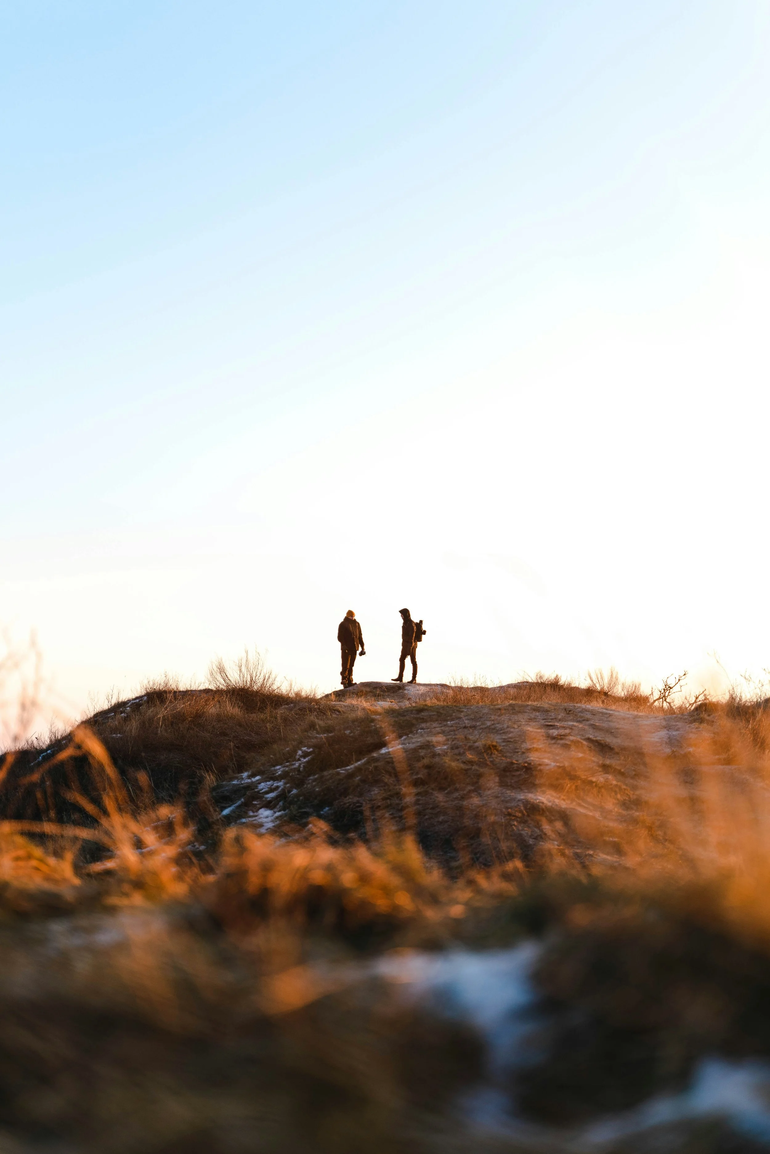 Two hikers standing on a hilltop at sunset or sunrise with a clear sky.