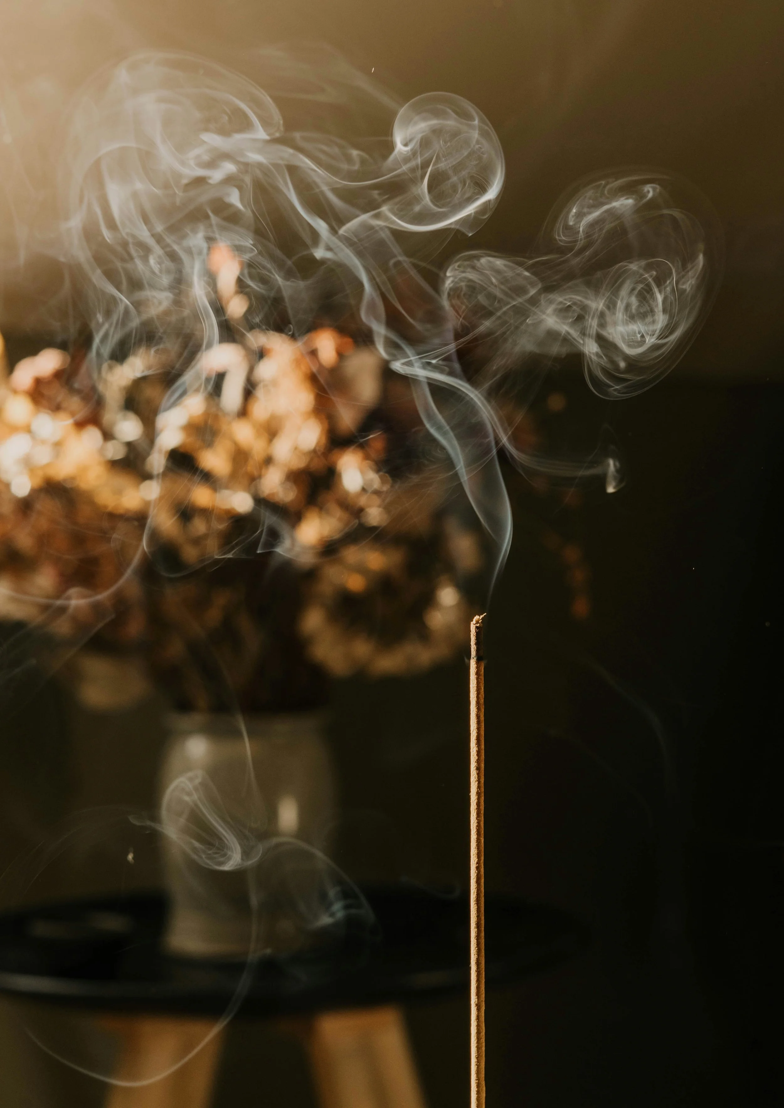 A lit incense stick with smoke curling upward against a dark background, with a blurred floral arrangement in the background.