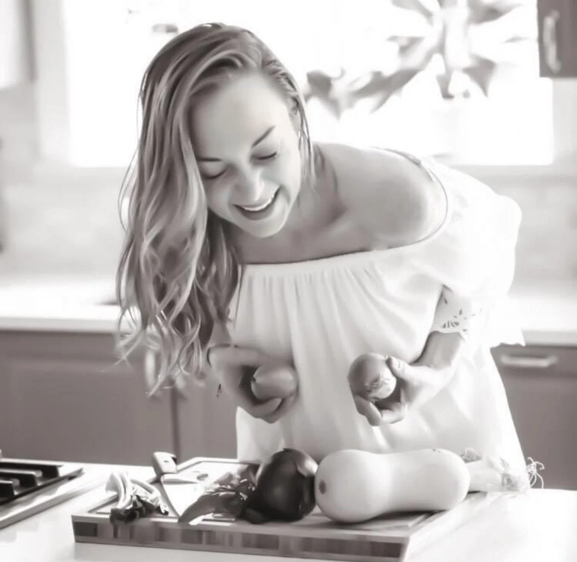 A woman with long, wavy hair, wearing an off-the-shoulder dress, is smiling while holding eggplants in each hand. There are more eggplants and a butternut squash on a cutting board in front of her in a kitchen.