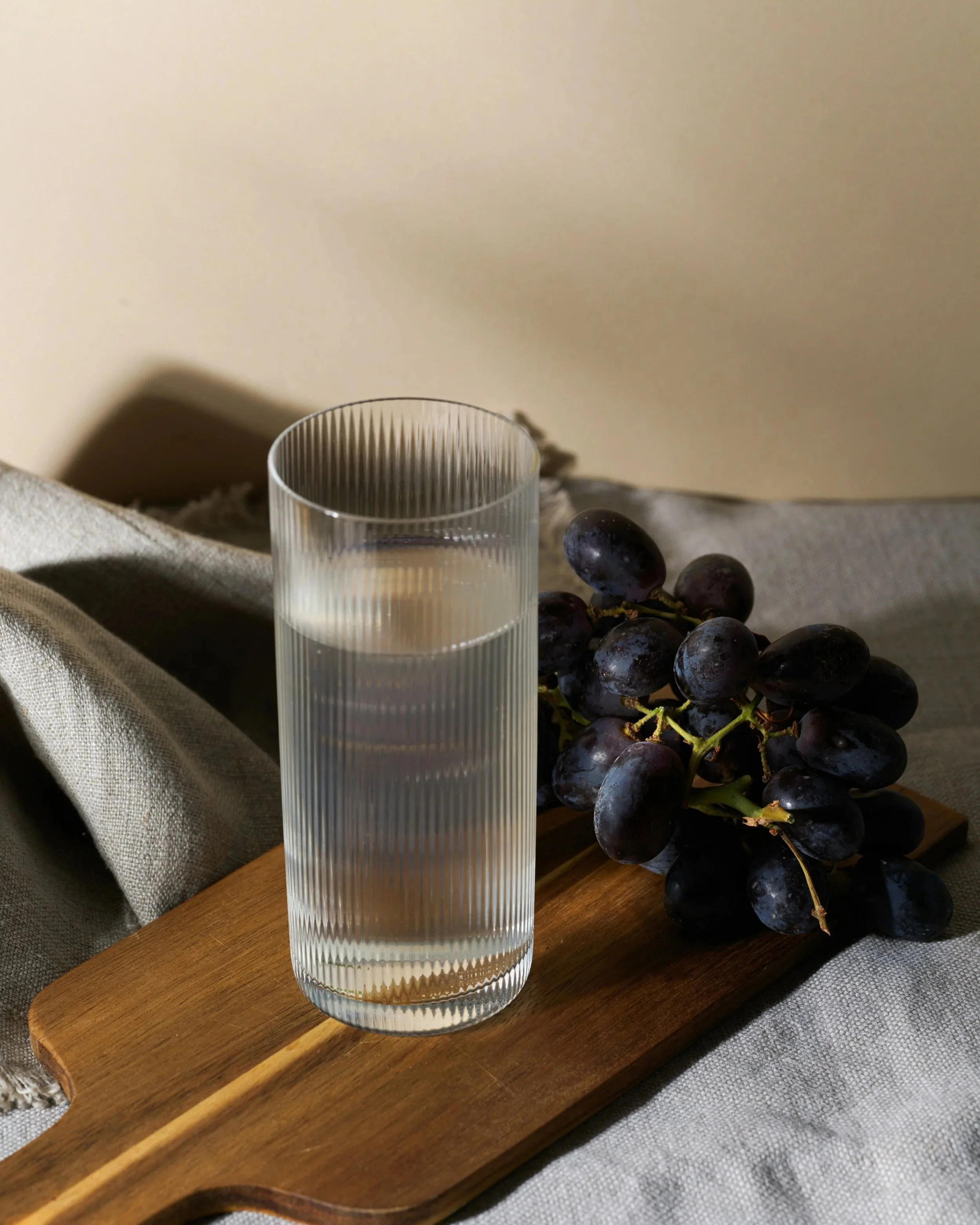 A tall, clear glass of water placed on a wooden cutting board, with a bunch of dark grapes beside it, on a textured fabric surface.