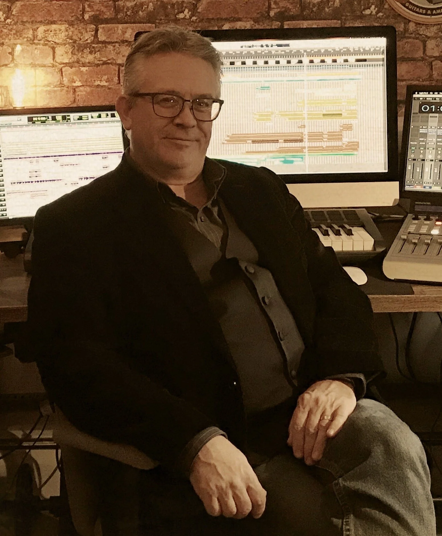 A man sitting in front of multiple computer screens with music production software and a keyboard in a studio with brick walls.