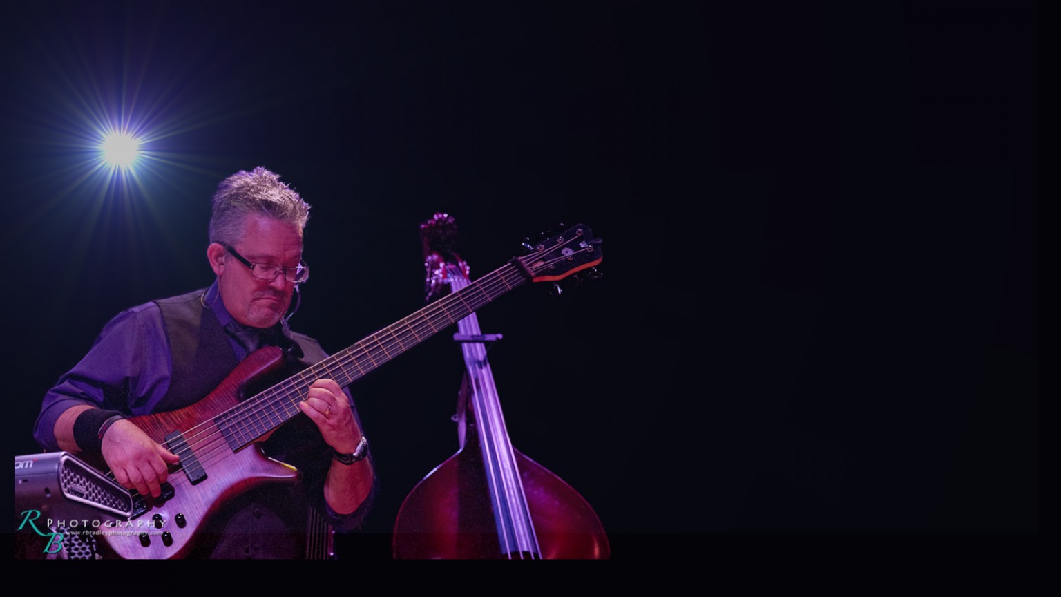 A male musician playing an electric guitar on stage with a double bass behind him, under purple lighting and a bright star-shaped light in the background.