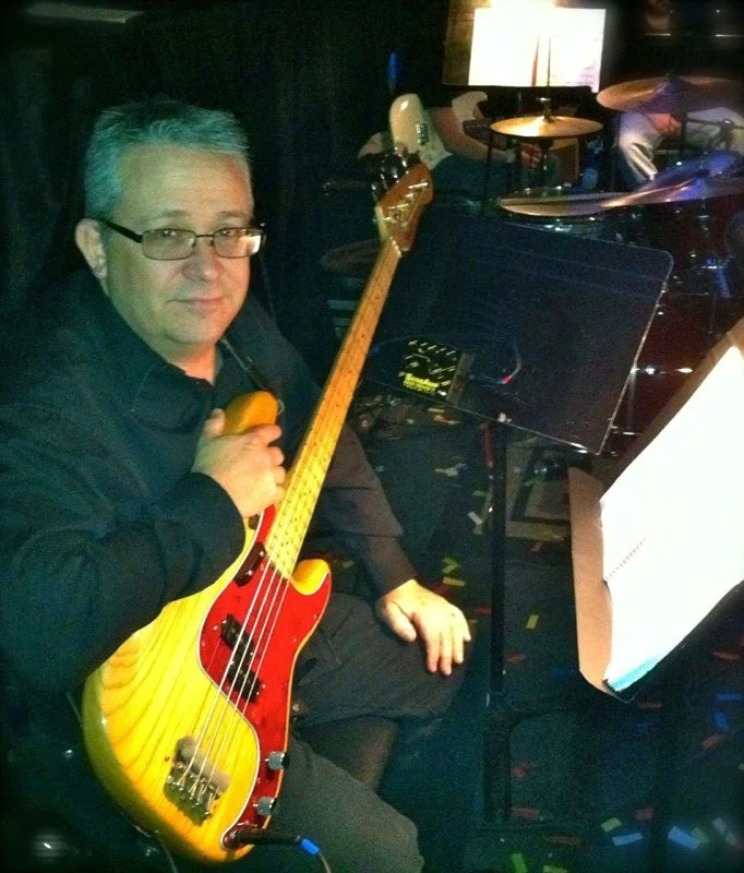 Man with glasses holding a yellow and red electric bass guitar, sitting in a music venue with music stands and drum set in the background.