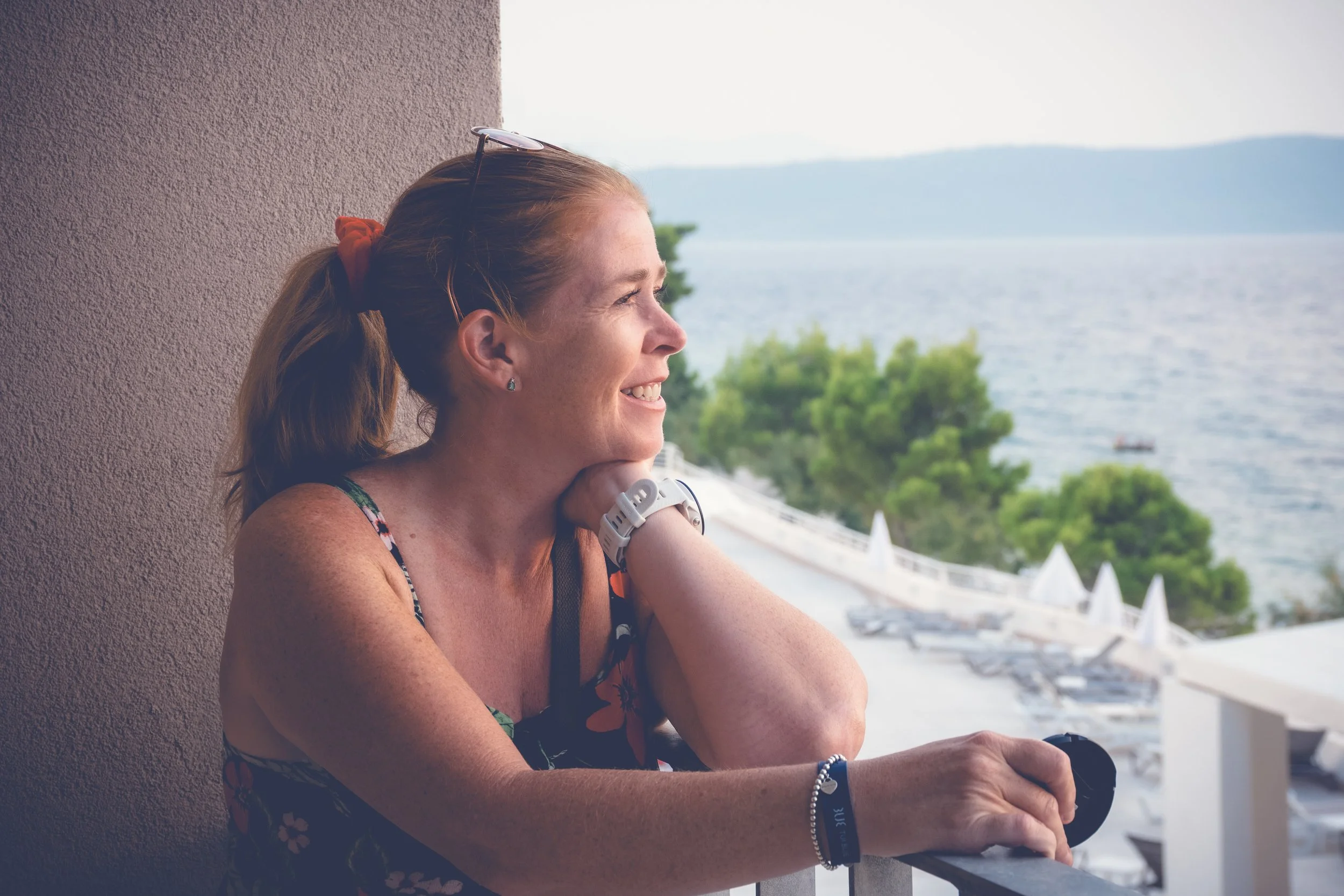 A woman with red hair tied back, wearing a floral tank top, smiling and looking out over a balcony with a view of the ocean, palm trees, and a beach with umbrellas.