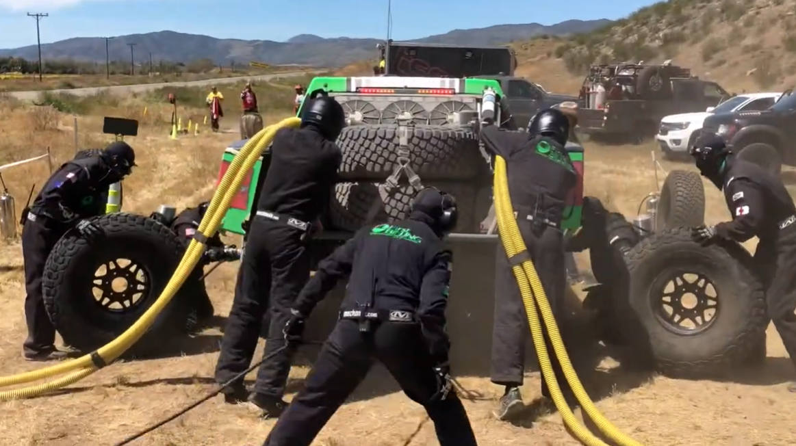 Race car crew members in black uniforms and helmets change a tire during a race on a dirt track with mountains in the background.