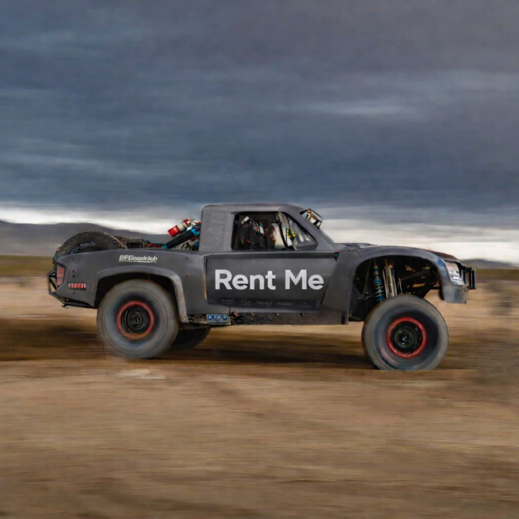 A modified off-road truck with a black body, racing tires, and a sign on the side that says 'Rent Me' driving on a dirt track under a cloudy sky.