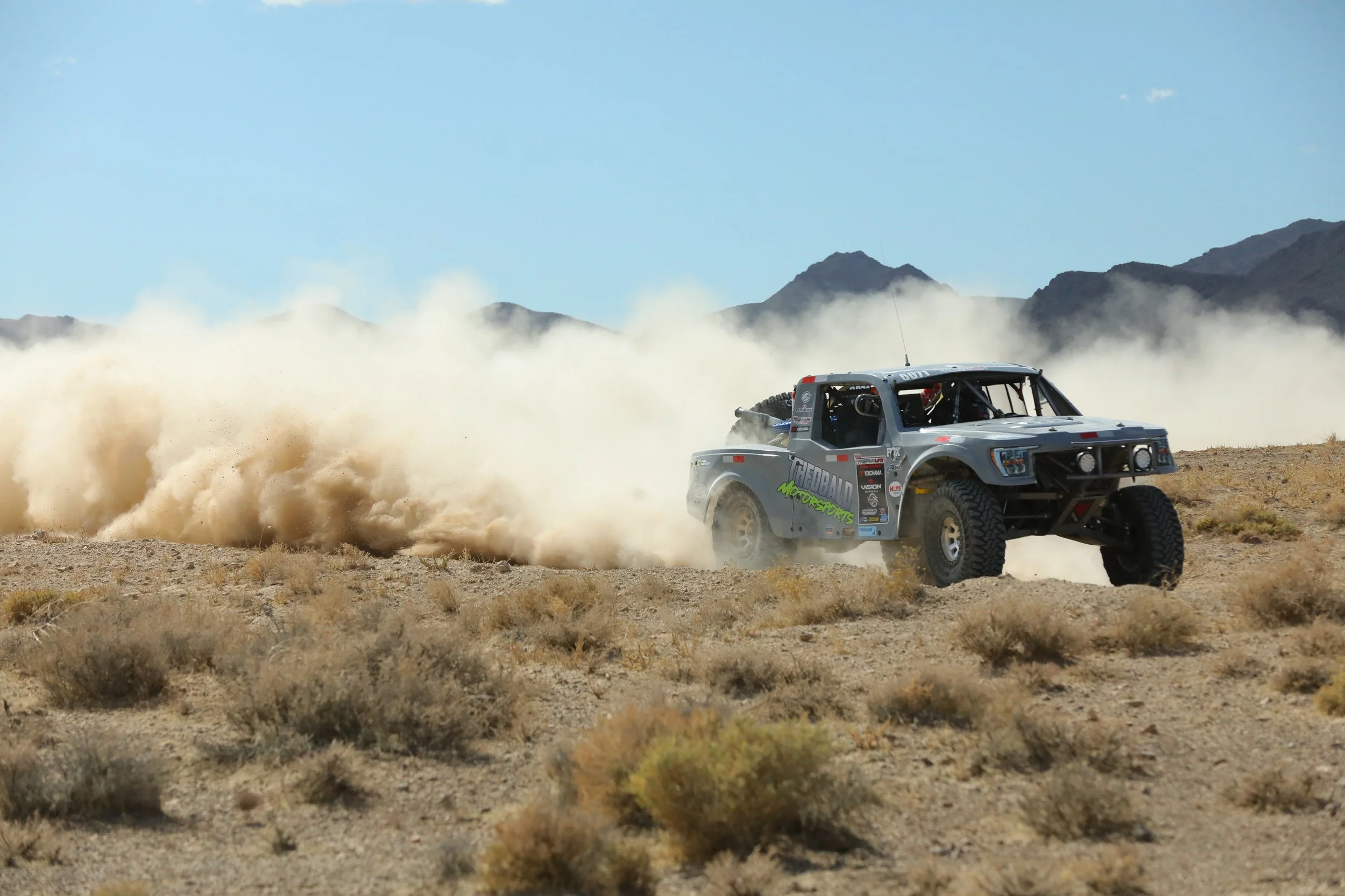 An off-road rally truck driving through desert terrain, kicking up a large cloud of dust with mountains in the background under a clear blue sky.