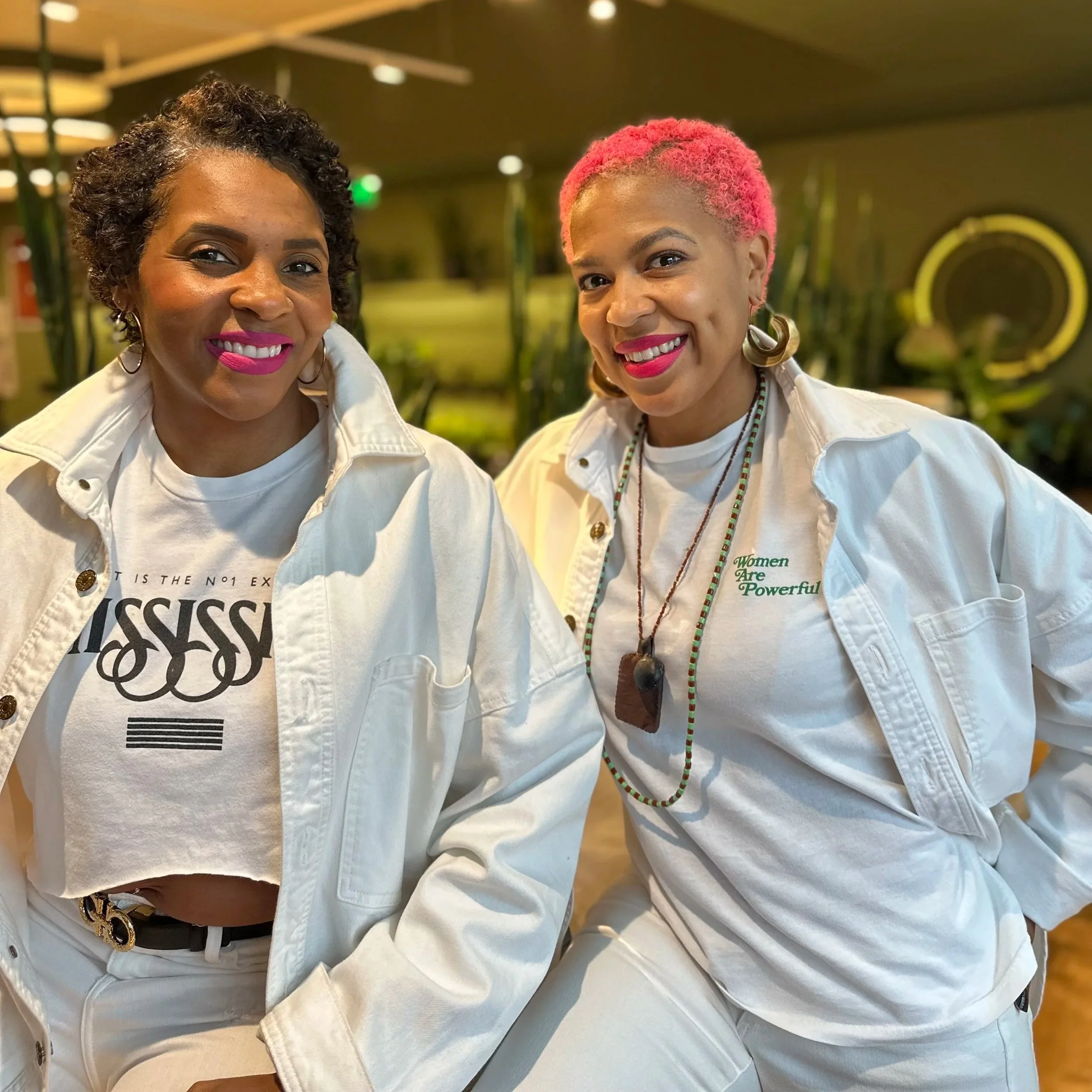 Two women with short hair and bright lipstick smiling, wearing white jackets and T-shirts, standing in a modern indoor setting with plants and warm lighting.