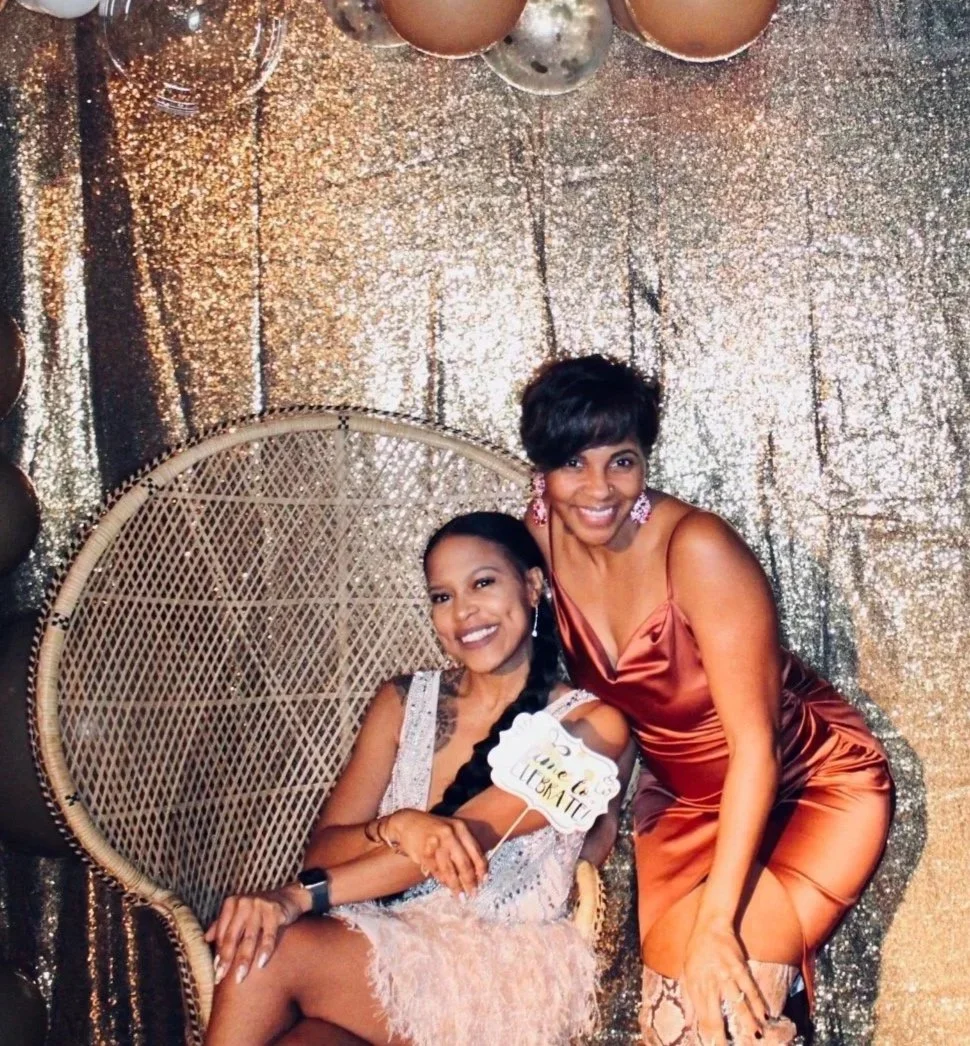 Two women posing and smiling at a party, one sitting in a wicker chair and the other standing beside her. They are dressed in elegant dresses with a glittery gold backdrop and balloons behind them.