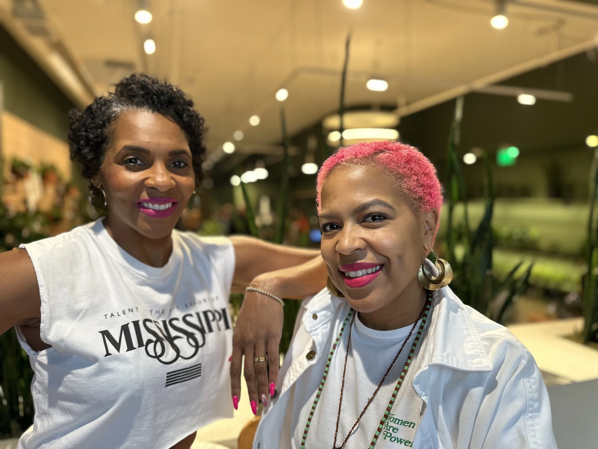 Two women smiling at the camera in a well-lit indoor space with plants in the background. The woman on the left has short black curly hair, bright pink lipstick, and is wearing a white t-shirt with black text. The woman on the right has short pink curly hair, earrings, and is wearing a white jacket with necklaces.