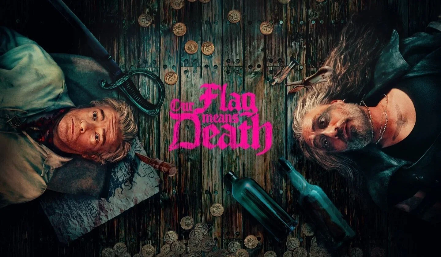 Two men lying on a wooden floor surrounded by coins, a bottle, a sword, and arrows, with the text 'Our Flag Means Death' in pink in the center.