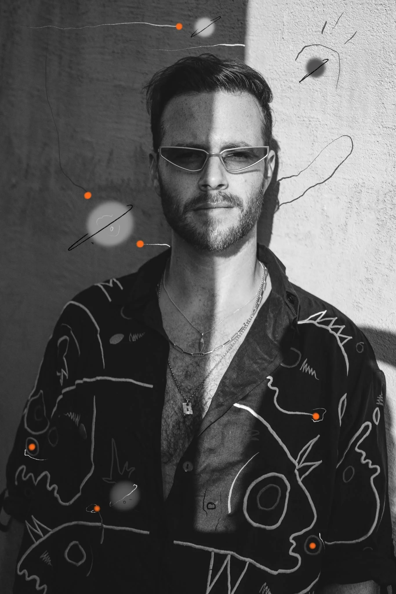 Black and white photo of a young man with styled hair, beard, wearing sunglasses, layered necklaces, and a patterned shirt, standing against a textured wall with line drawings and small orange dots overlayed.