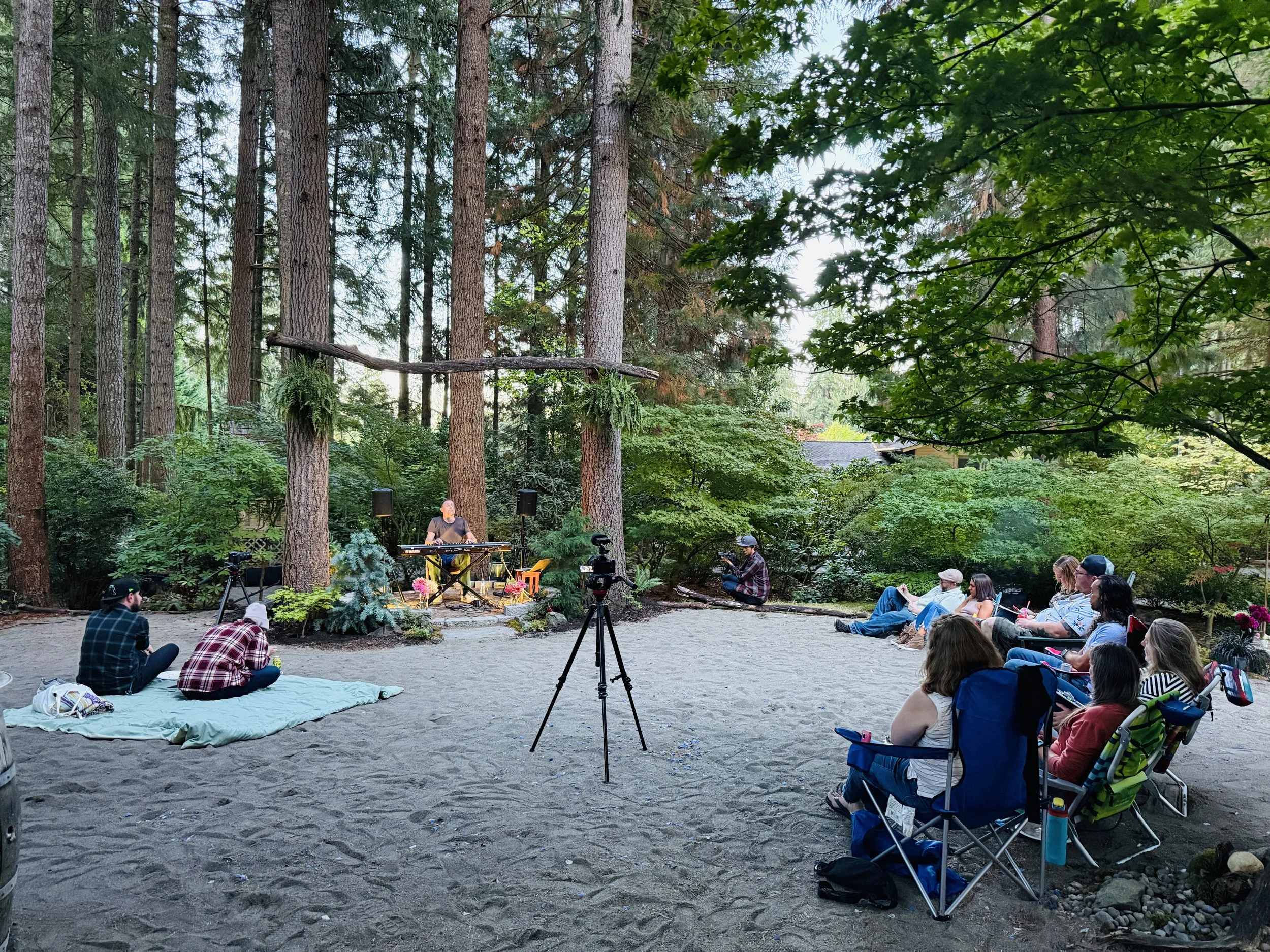 People sitting on chairs and blankets in a forested outdoor area, attending a live music performance with a keyboard player on a small stage, surrounded by tall trees and greenery.