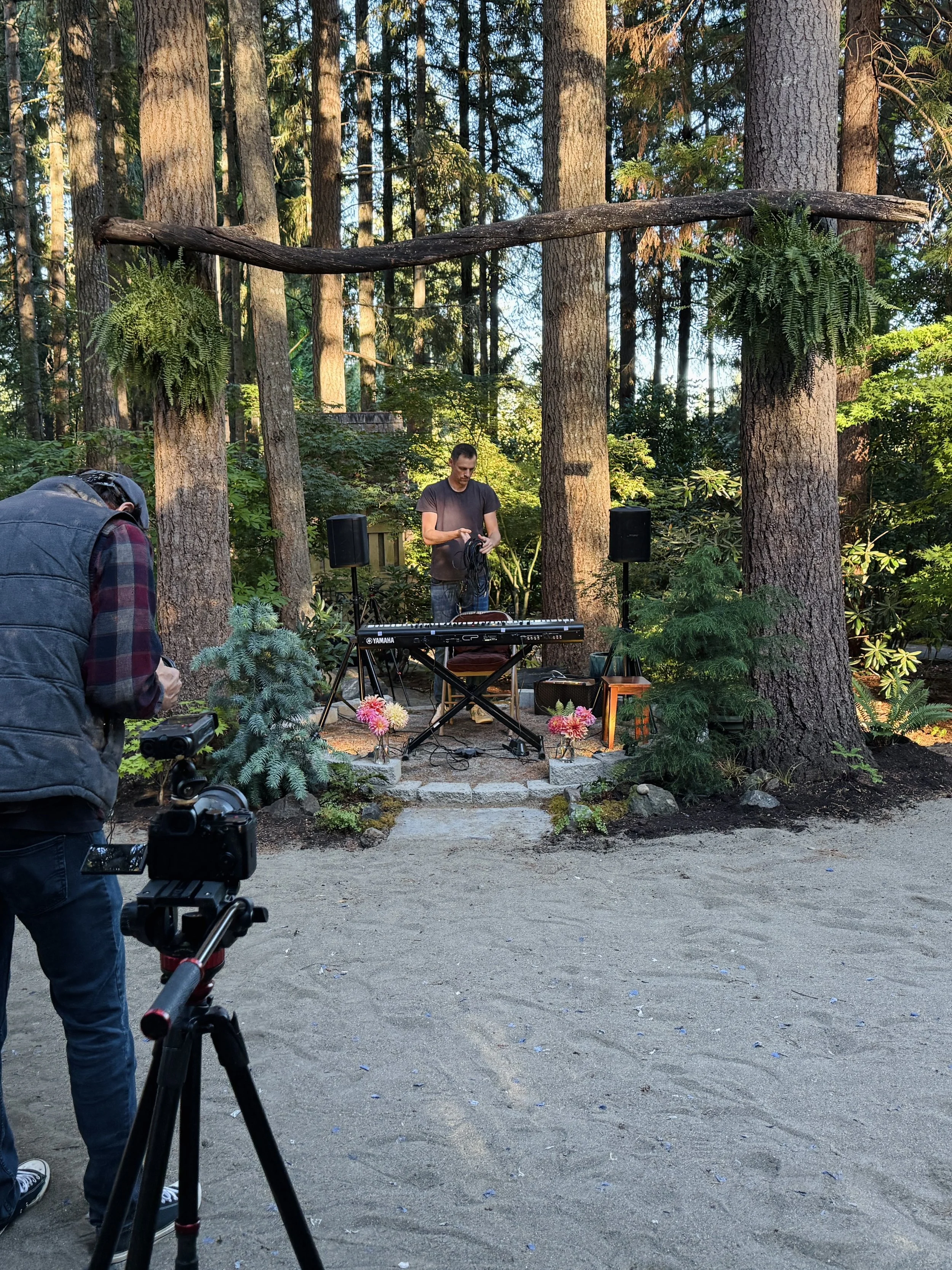 A young man stands behind a keyboard setup in a wooded outdoor area during daytime, with a photographer and camera in the foreground capturing the scene. There are potted plants and flowers around the stage area, and large trees with hanging ferns ov