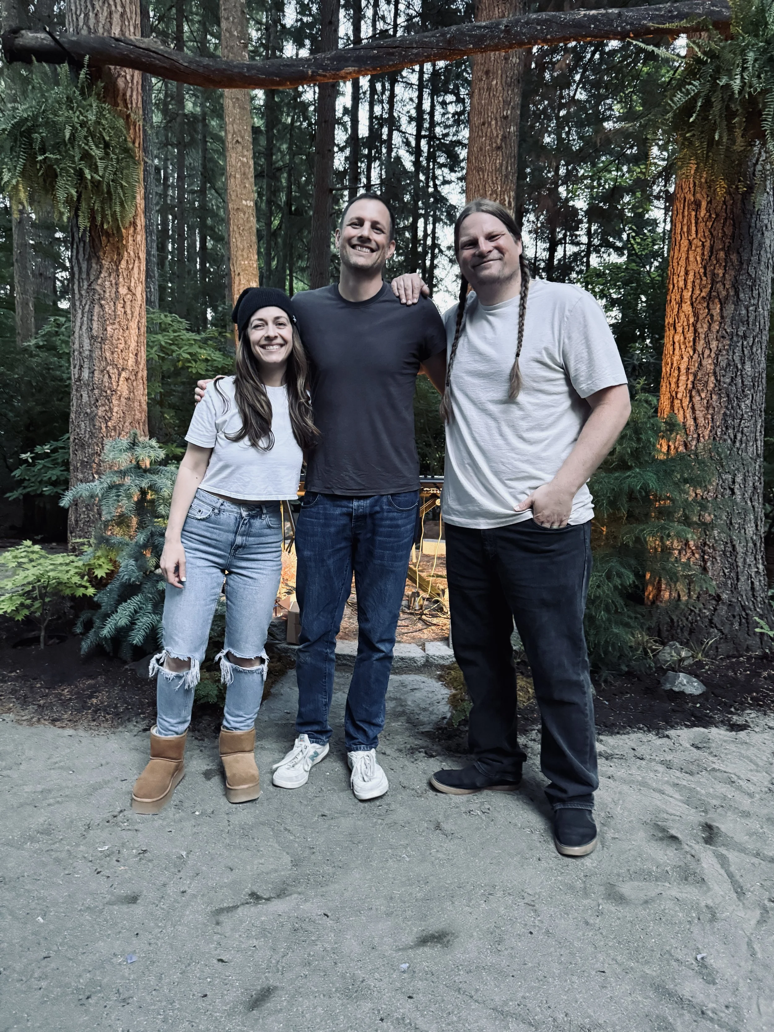 Three friends smiling and standing outdoors in a forest, posing for a photo with trees and greenery in the background.