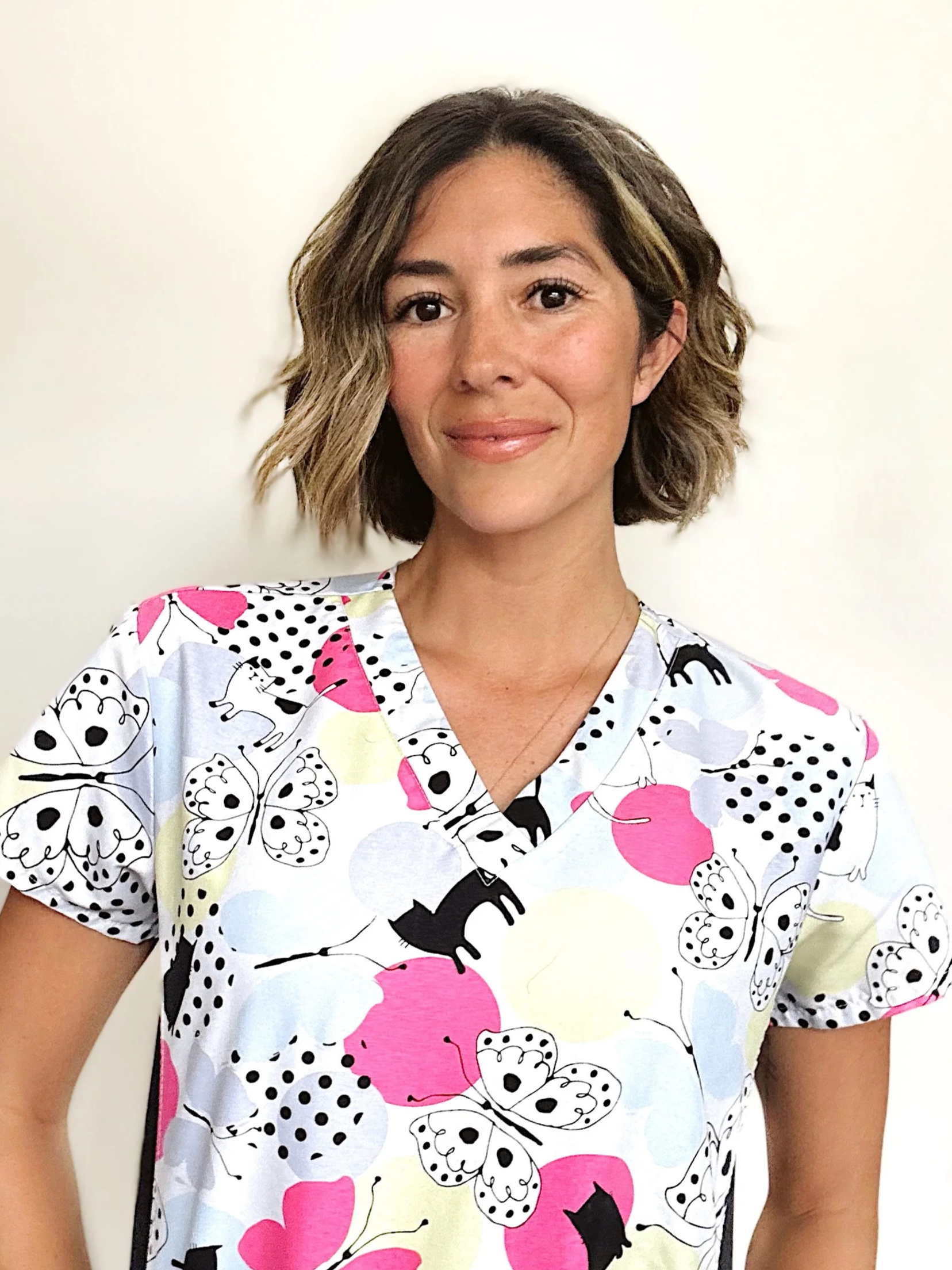 A woman with short wavy brown hair and light skin, smiling, wearing a colorful scrub top with butterfly, cat, and polka dot patterns in pink, black, yellow, and light blue, against a plain white background.