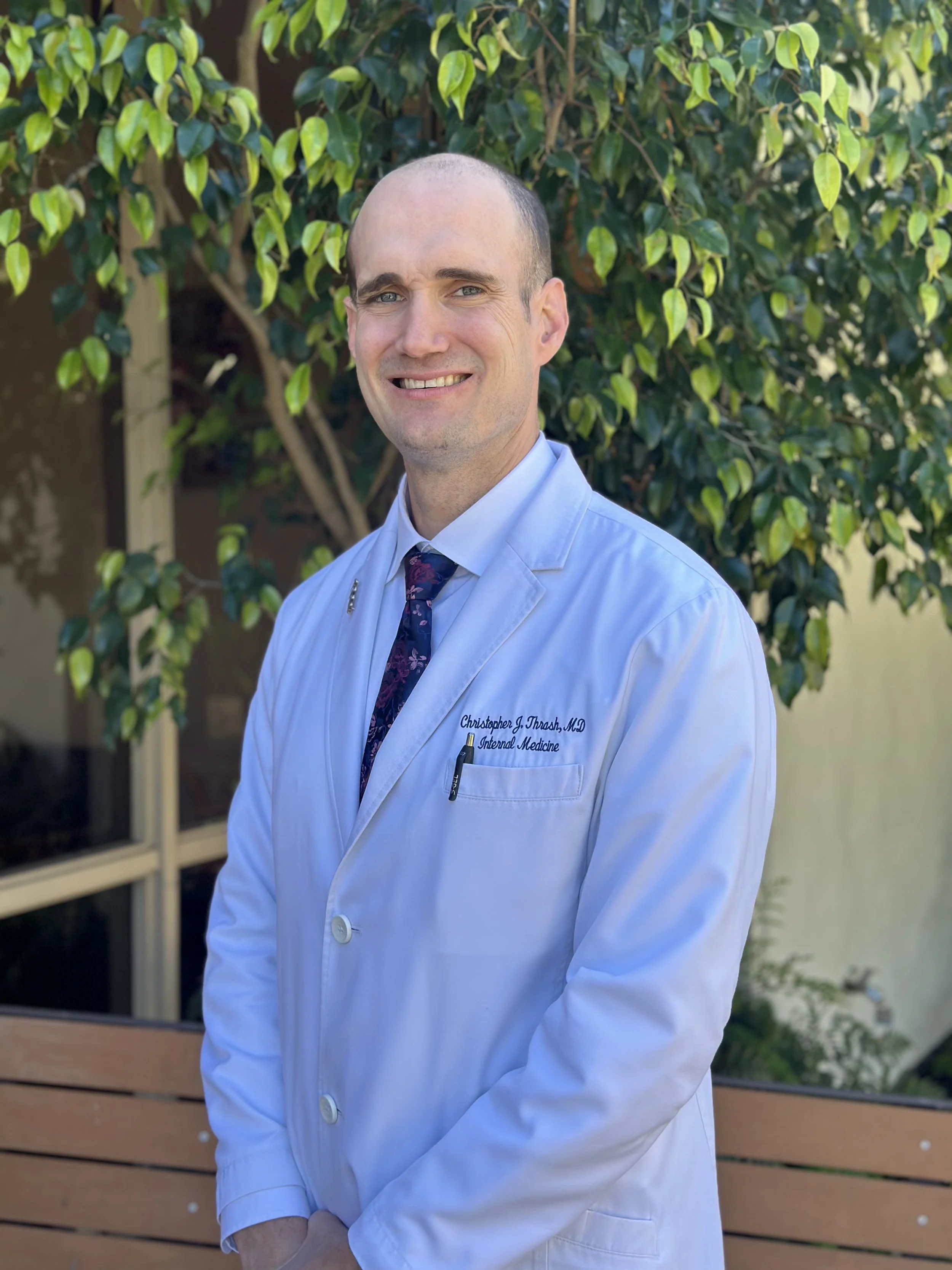 A male doctor standing outdoors in front of green foliage, wearing a white lab coat and a patterned tie, smiling at the camera.