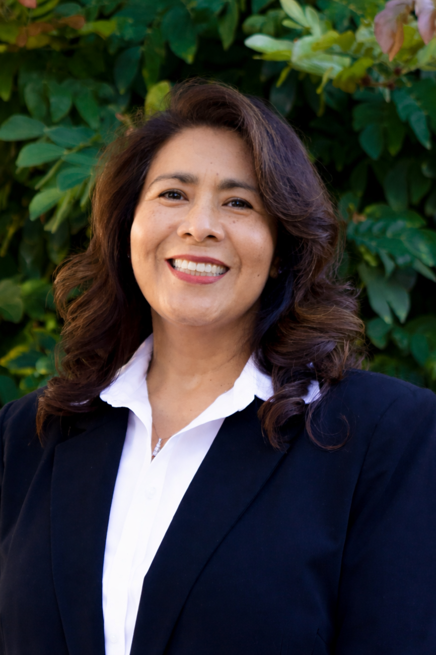 A smiling woman with wavy brown hair, wearing a black blazer and white shirt, standing outdoors in front of green foliage.