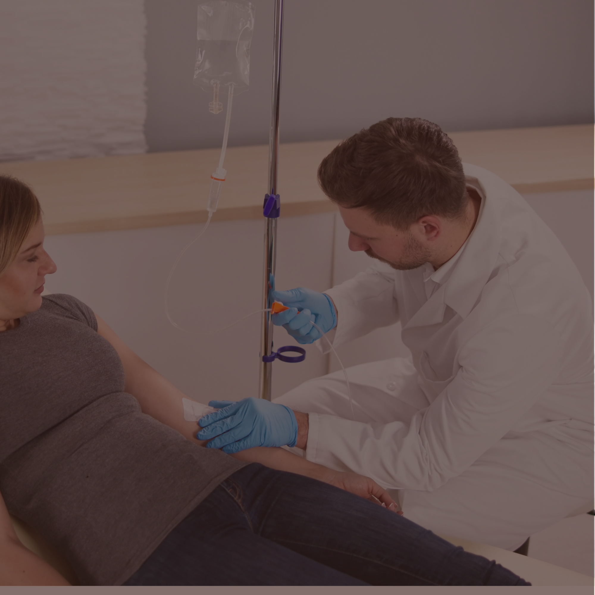A healthcare professional in white coat and blue gloves is administering an intravenous (IV) infusion to a patient lying on a bed. The IV bag is hanging from a stand, connected to a tube that leads to the patient's arm.