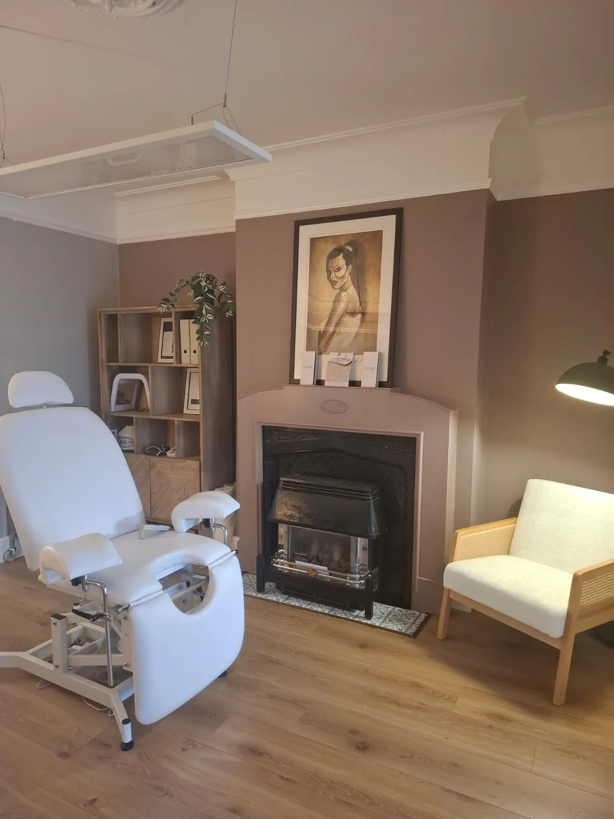 Medical examination room with a white reclining chair, a fireplace with artwork above, a bookshelf with various items, and a small beige armchair with a lamp nearby. This is the main room of a wellness, healthcare clinic.