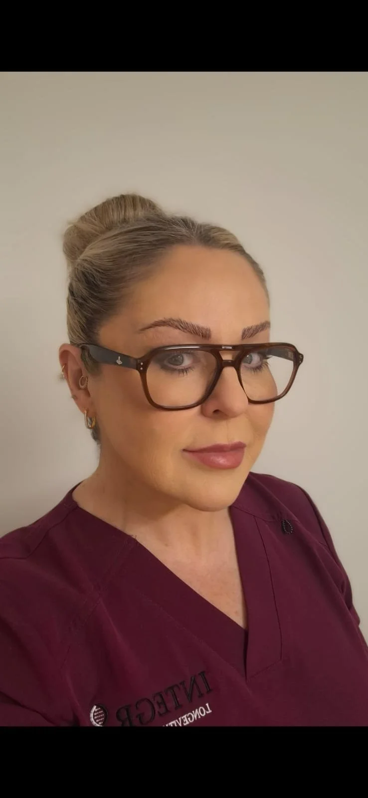 A qualified phlebotomist and nursing assistant. A headshot of a woman with blonde hair in a bun, wearing large glasses and a maroon medical uniform, standing against a plain wall in her work scrubs.