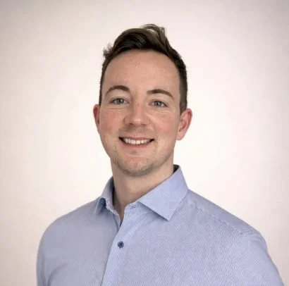 A qualified chiropractor. the headshot shows a man with short dark hair, wearing a light blue collared shirt, smiling at the camera against a plain light background.