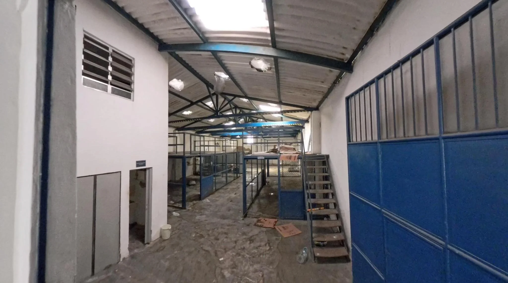 An empty industrial-style interior with a corrugated metal roof and exposed steel beams, featuring blue-framed partitioned bays, a narrow metal staircase to a small platform, and a dusty floor with scattered debris and cardboard.