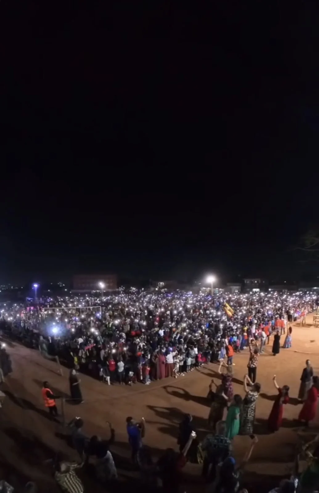 A massive nighttime crowd in an open field, lit by thousands of phone lights.