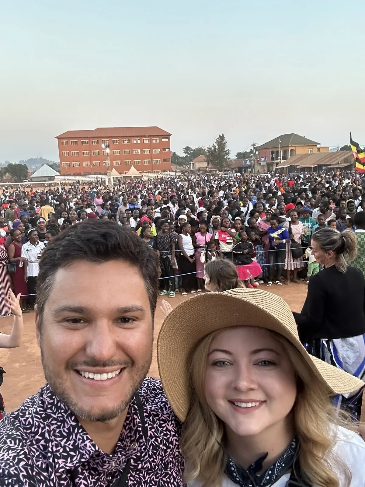 A smiling man and woman (the woman wearing a wide-brim straw hat) take a selfie at an outdoor event, with a large crowd gathered behind a rope barrier and buildings and a flag visible in the background.