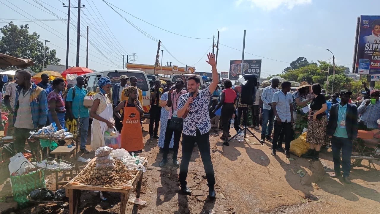 A man in a patterned shirt stands on a dusty roadside holding a microphone and raising one hand as he speaks, surrounded by a crowd near a market stall with food on a table, a parked van, and overhead power lines in bright daytime.